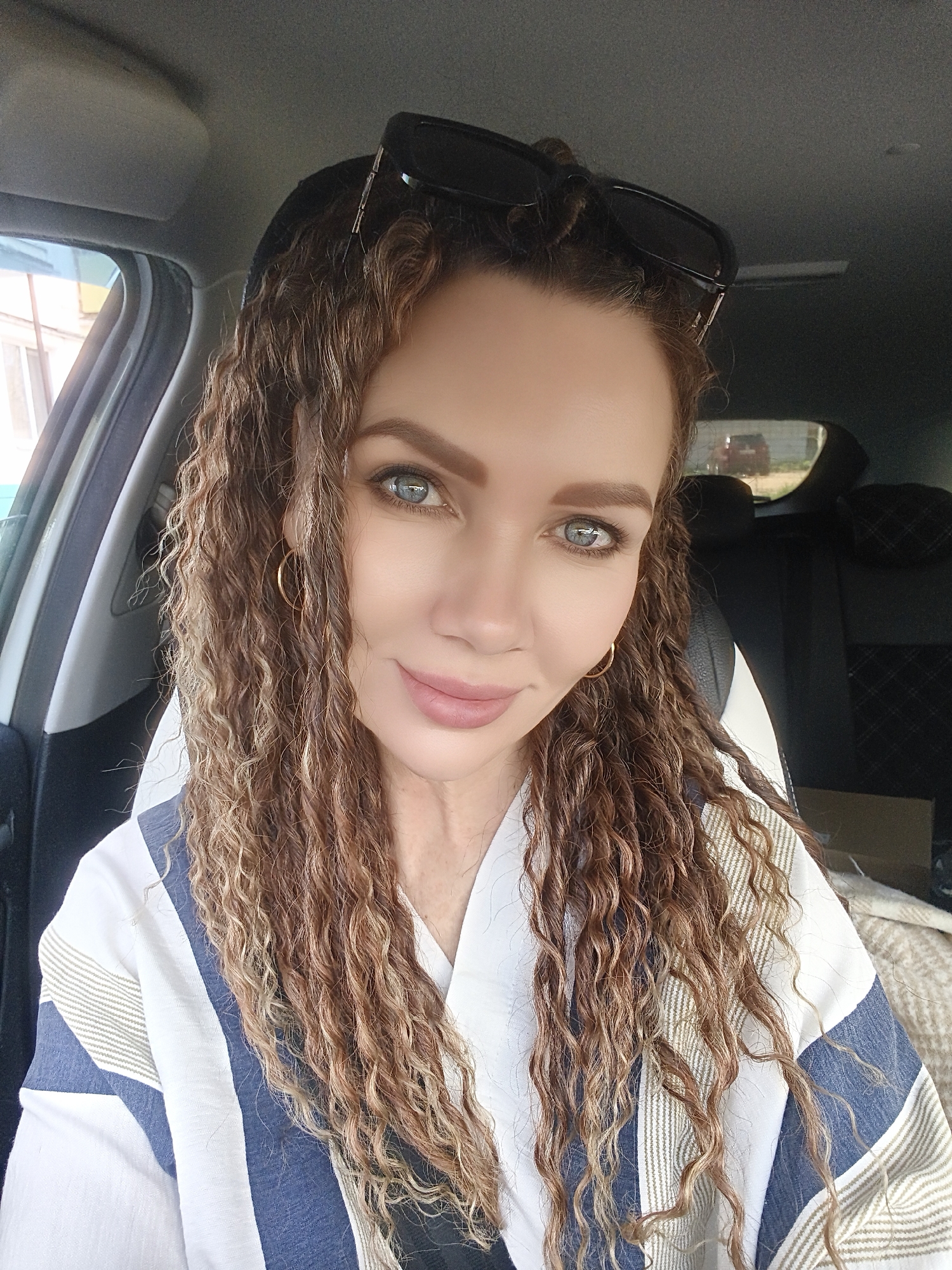 A woman with curly brown hair is in a car. She is wearing a white and blue striped shirt and gold hoop earrings. Sunglasses are perched on her head. The car's interior includes black seats and a white headrest. Through the car window, a building can be seen.