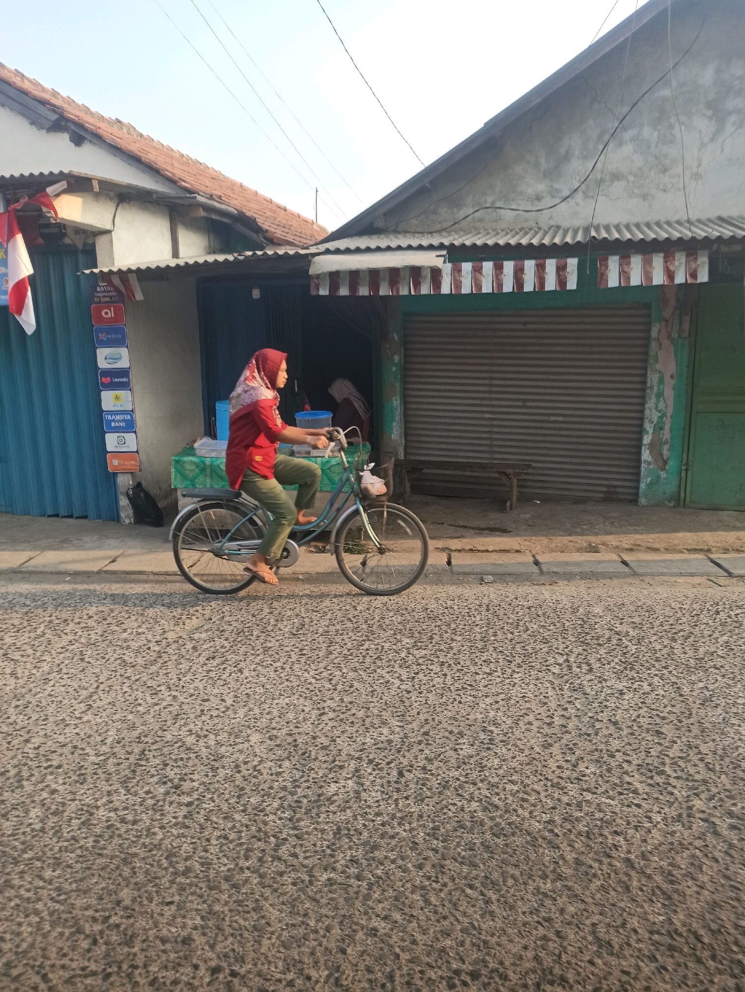 A woman wearing a red shirt and green pants, with a red and white headscarf, is riding a bicycle on a paved street in front of a building. The building appears to be a shop or a series of shops with corrugated metal roll-up doors and awnings. There are several signs visible on the left side of the building, including one that says "TRANSFER BANK" and another with the Intel logo. A small wooden bench is placed in front of the closed shop. The scene is set in Telagamurni, Indonesia, and the time of day appears to be late afternoon or early evening due to the warm lighting. The street is made of asphalt with visible cracks and a concrete curb.