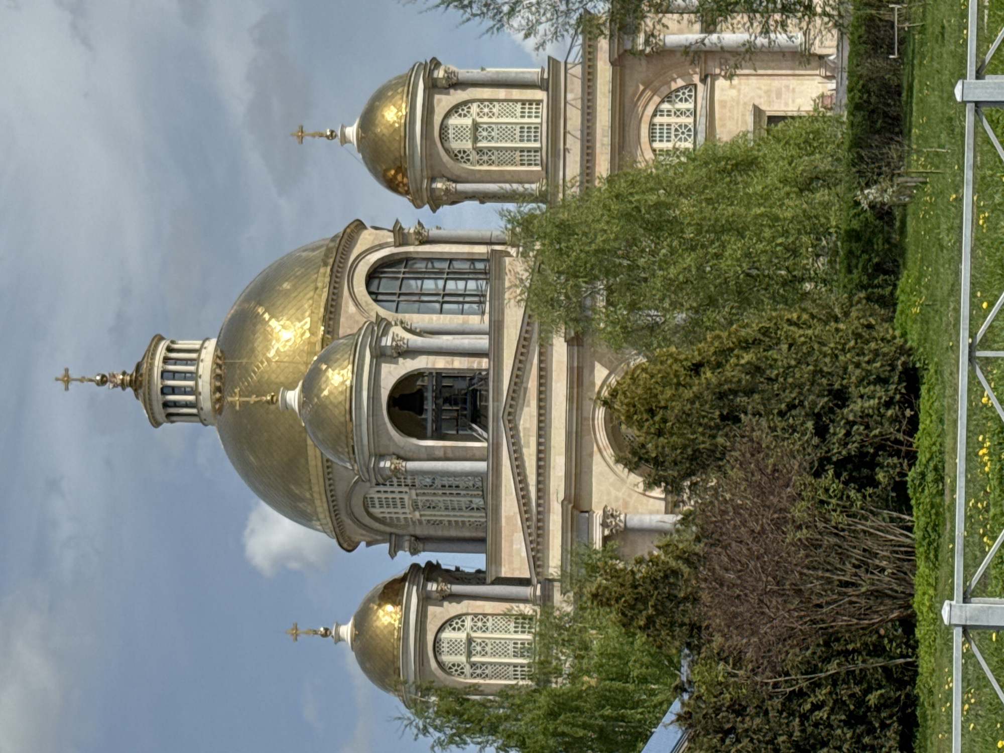 Captured from a ground-level vantage point, the photograph showcases the architectural grandeur of a Russian Orthodox church, likely situated in Saint Petersburg. The church boasts several golden domes that glisten brilliantly under the sun's illumination, punctuated with intricate architectural details such as windows and embellishments. A lush canopy of trees partially obscures the view of the church, introducing a natural element into the urban landscape. A metallic fence post, located at the right edge of the frame, indicates the boundary between the architectural structure and the surrounding natural environment. The sky, partially visible, adds depth to the composition.