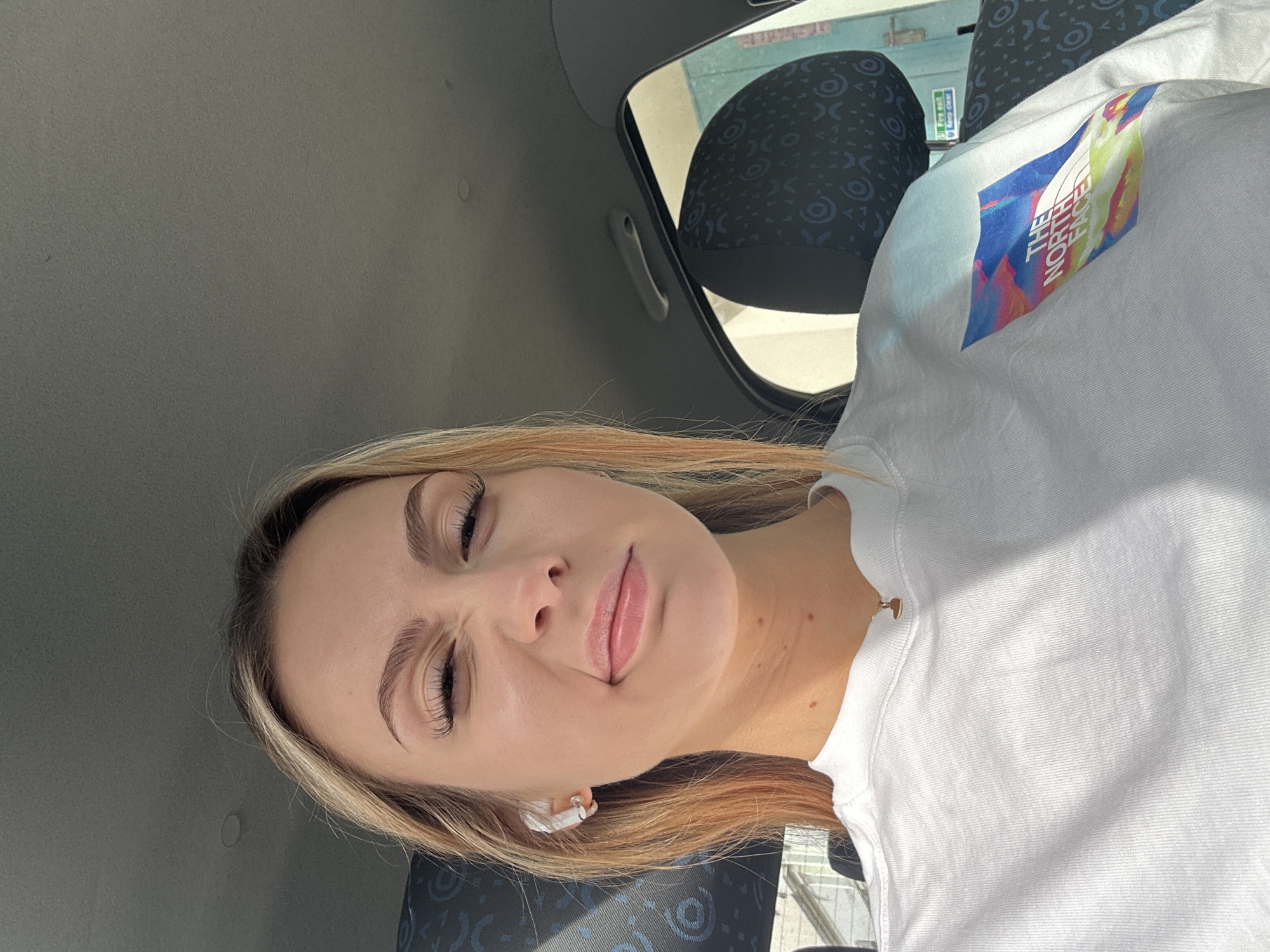 A portrait-style photograph shows a blonde woman wearing a white T-shirt featuring a logo that includes the text "THE NORTH FACE". She is seated in what appears to be a vehicle interior, based on the visible car seat fabric and interior panels. The location is noted as the city of Unknown, Unknown.