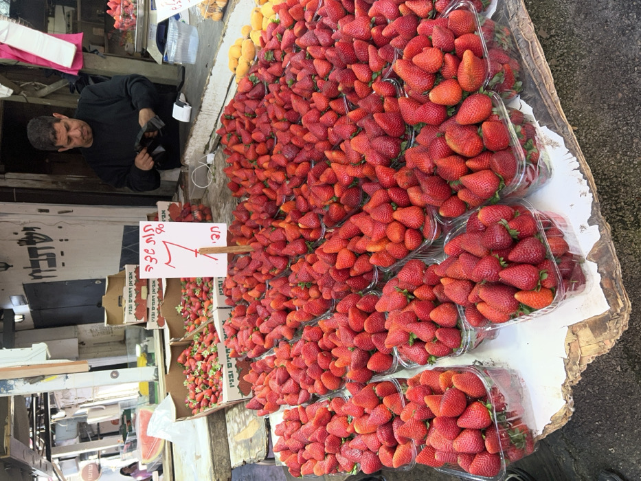 This image depicts a bustling produce stall within an outdoor market, consistent with the environment of Shuk HaCarmel in Tel-Aviv, Israel. The foreground and midground are dominated by an extensive display of fresh, ripe, red strawberries, meticulously arranged in numerous clear plastic containers on a long, weathered wooden counter with a peeling white surface.
A prominent white sign with red Hebrew text is centrally placed among the strawberries, reading "תות שדה" (Strawberry), "חבילה" (Package/Box), and the number "7", likely indicating a price. Behind the counter, an adult male vendor with dark hair and a beard, wearing a dark long-sleeved shirt, is observed looking downwards. He is actively engaged, manipulating a small, dark object with both hands over a white thermal receipt printer positioned on the counter.
In the background, stacked cardboard boxes contain additional strawberries; some boxes bear the Hebrew inscription "אביא רחמים ובני" (Avia Rachamim and Sons) and "ישראל" (Israel). To the far left, a partial slice of watermelon rests on a white surface, while a small pile of yellow fruits is visible on the far right of the main strawberry display. The market setting includes an open-air structure with visible overhead metal framework, older building facades with faded paint, and distant, blurred market activity. Natural light illuminates the scene.