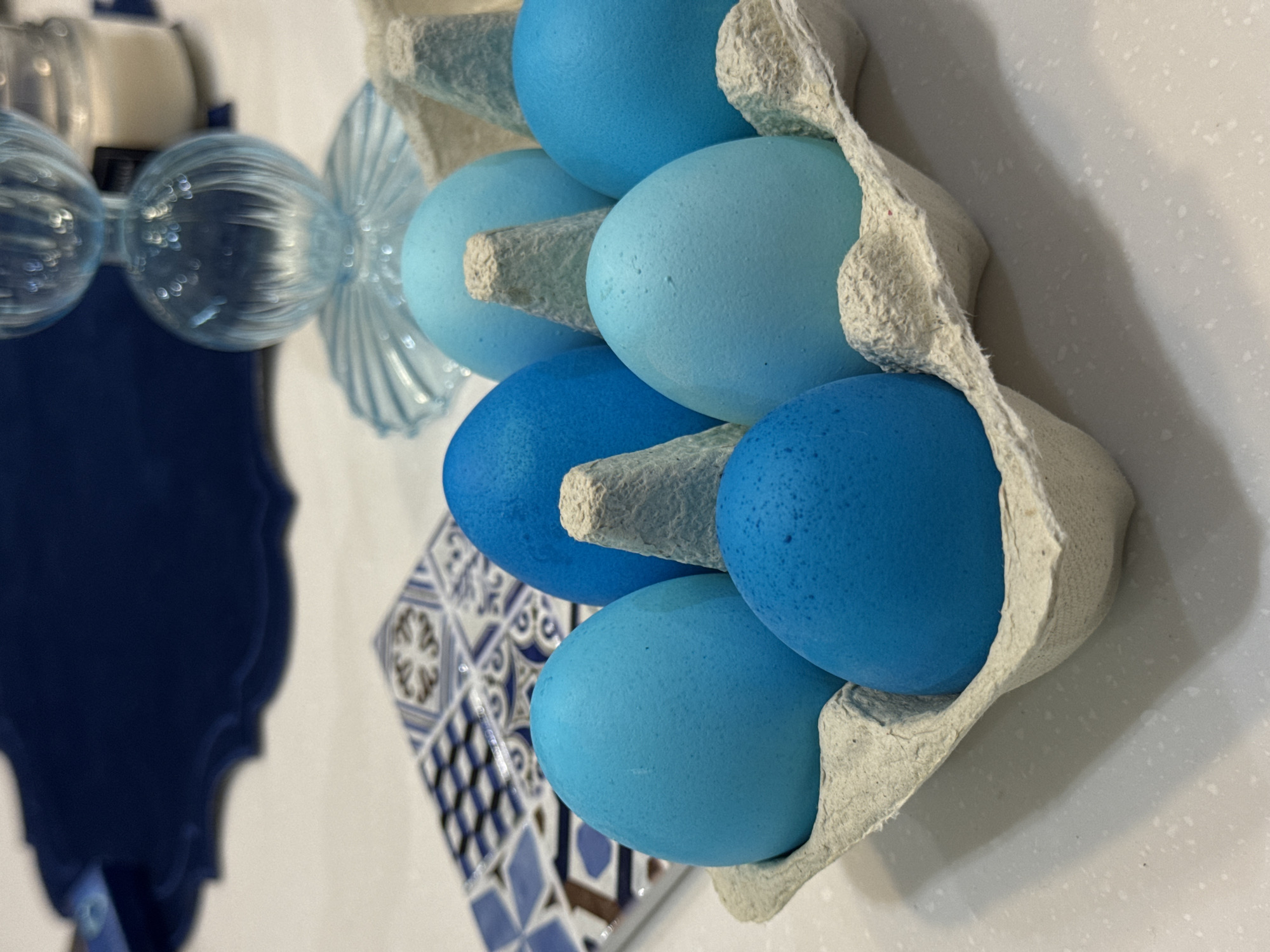 A close-up shot shows a carton of six blue-dyed eggs sitting on a white countertop, partially obscuring a blue and white patterned tile and a blue decorative object with glass elements. The eggs vary in shade, from a lighter pastel blue to a deeper azure. The egg carton is a light gray, textured paper material. The decorative object has a dark blue backing and a lighter blue, glass front with curved elements. The photo appears to have been taken indoors. There is no information to suggest that the location of this image is in Khimki, Russia.