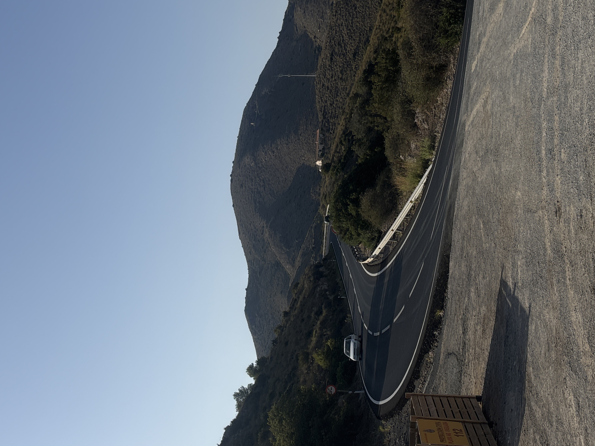 The image shows a winding asphalt road cutting through a hilly, arid landscape under a clear blue sky. A white sedan is traveling on the road, following its curves. The roadside is lined with sparse vegetation, including bushes and small trees. A metal guardrail runs along the outer edge of the road. In the background, a large, undulating hill dominates the scene, with a few scattered buildings and a cell tower visible on its slopes. In the foreground, a wooden barrier with yellow signage is present, indicating "PROTECCIÓN CIVIL PLAYA NO VIGILADA 112" which translates to "Civil Protection Unsupervised Beach 112". This suggests the location is near a coastal area in La Unión, Spain, specifically indicating emergency services information for an unsupervised beach. The overall impression is one of a rural, scenic route.