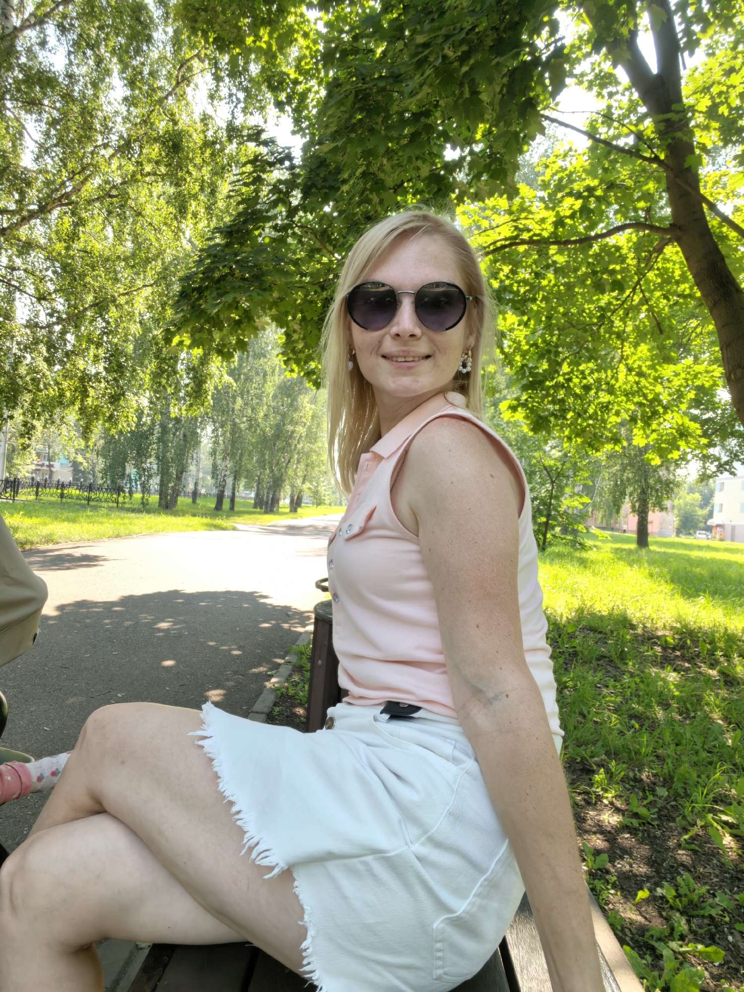 A woman with blonde hair and sunglasses is sitting on a park bench, with trees and a path behind her. The location is the city of Sterlitamak, Russia.