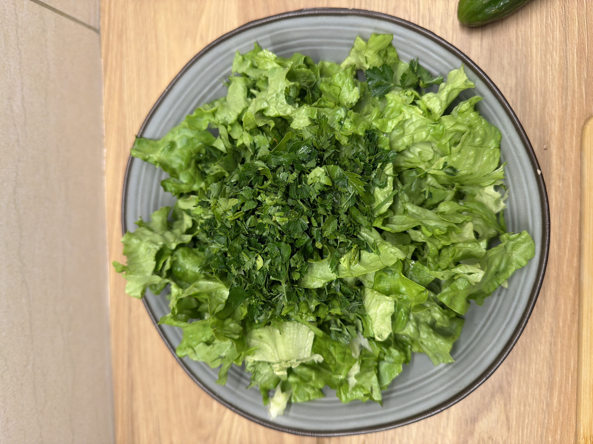 A top-down shot captures a fresh green salad prepared in a round, grey bowl with a dark brown rim. The bowl is primarily filled with vibrant green lettuce leaves, which appear freshly chopped and slightly moist. Centered on top of the lettuce is a mound of finely chopped dark green herbs, likely parsley or dill.
The bowl is situated on a light-colored wooden surface, possibly a cutting board or kitchen table. A small, dark green cucumber is partially visible in the upper right corner of the frame, resting on the same wooden surface, hinting at additional ingredients or preparation. To the left, a light beige, textured surface, perhaps a countertop or wall, is visible. The scene suggests an indoor setting, likely a kitchen or dining area, focusing on the preparation of a healthy meal. There is no visible text or explicit location information within the image itself.