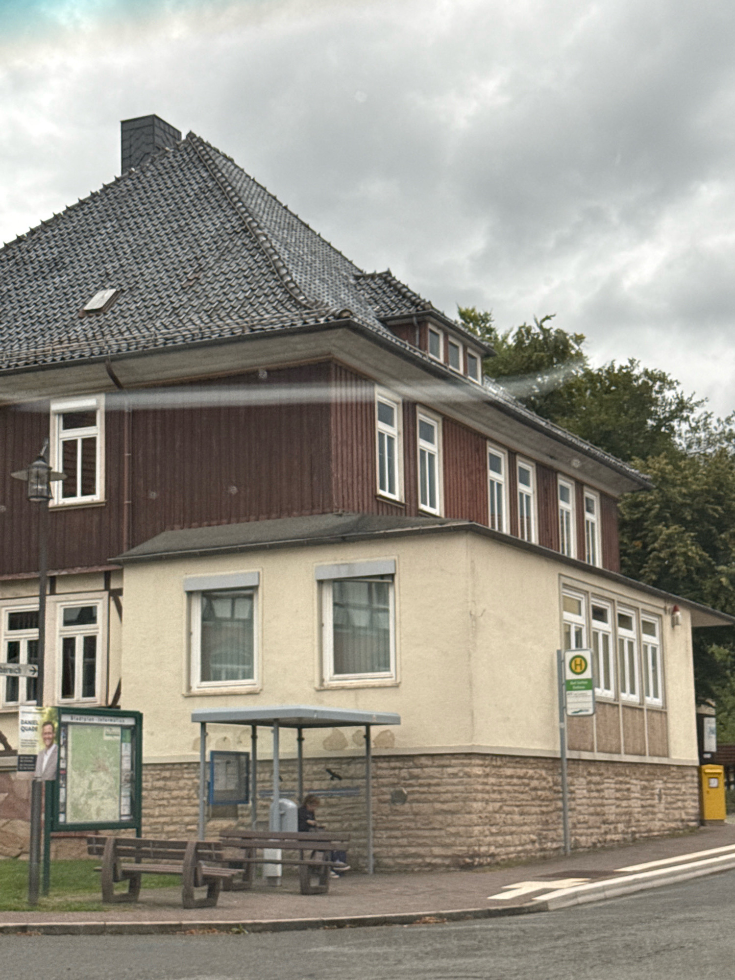 The image shows a building exterior in Bad Sachsa, Germany, with a bus stop in the foreground. The building is a two-story structure with a dark grey tiled roof and dark brown wooden siding on the upper level. The lower level is cream-colored with a stone base. Multiple windows are visible on both levels.
At the bus stop, there are two wooden benches, a grey metal frame with a roof, and a display board with maps and information. A person is seated on the bench on the right, looking at their phone. A yellow mailbox is visible to the right of the bus stop. A street sign with a white "H" in a green circle indicates the bus stop. A poster with a man's face and text is attached to a pole to the left of the display board. The environment appears to be overcast with green trees in the background.