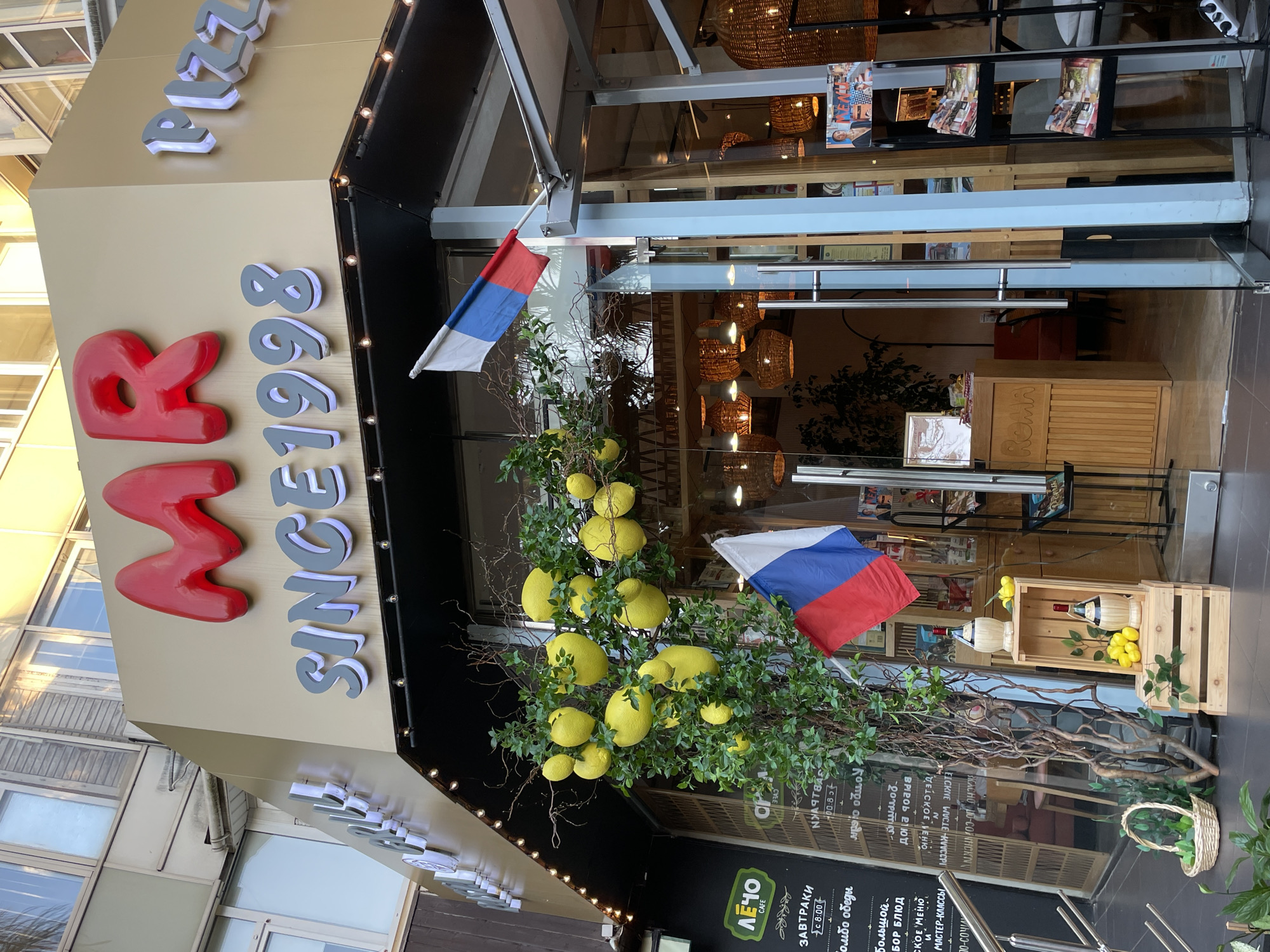 The image shows the exterior of a restaurant called "MR. PIZZA SINCE 1998" located in Sochi, Russia. The restaurant has a prominent red sign with the letters "MR" and "PIZZA" in white, illuminated letters. There are decorative elements such as a tree with large yellow lemons and two flags, one of which is the Russian flag. The entrance to the restaurant is visible, with glass doors and a display of what appears to be menus or promotional materials. The time of day appears to be late afternoon or early evening, as indicated by the warm lighting. There are no people visible in the image.