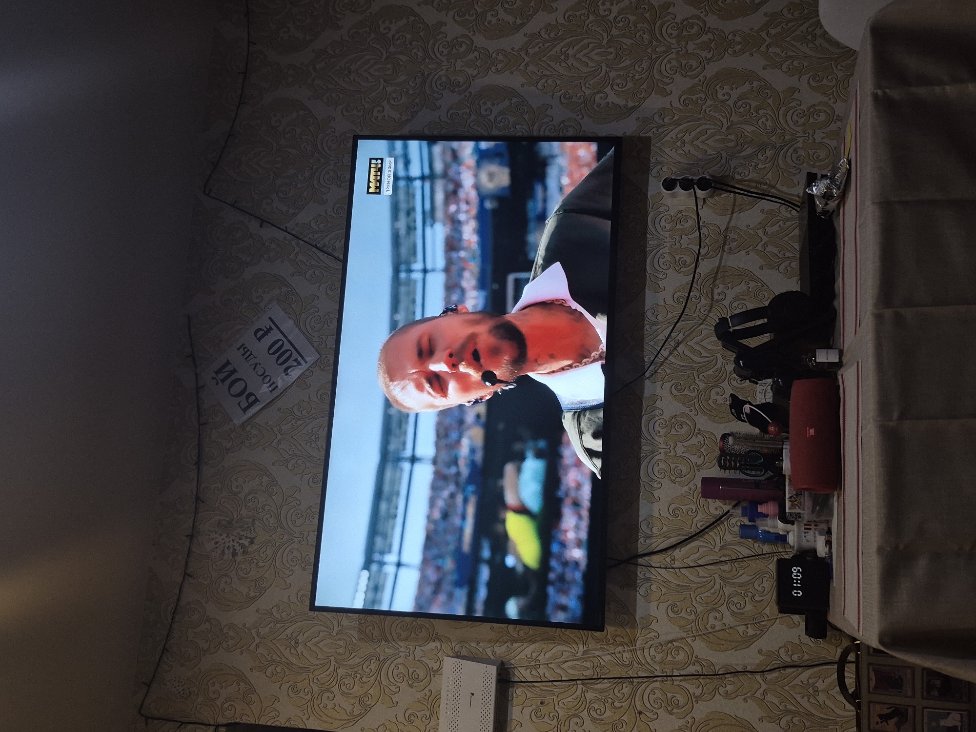 The image depicts a living room setting with a large flat-screen television dominating the central view. The television displays a performance by a male musician with facial tattoos, wearing a white collared shirt under a dark jacket, with a microphone near his mouth and earphones in his ears. Behind him, a blurred stadium crowd is visible, suggesting a live concert or sporting event broadcast.
In the upper right portion of the image, a poster is adhered to the patterned wallpaper, featuring Russian text and a price of "200 P". The wallpaper itself has a recurring damask pattern in a muted yellow and beige.
Below the television, a table is covered with a beige tablecloth adorned with red and white stripes. On the table, several objects are present: a black digital clock displaying "01:09", various bottles and containers of toiletries, a red JBL portable speaker, a pair of headphones, and what appears to be an electronic device. To the left of the table, a framed picture or set of pictures is visible.
The room appears to be an indoor residential space, likely a living room, based on the furniture and typical household items. The lighting suggests it is either evening or the room's ambient light is low.
The location of Chaikovsky, Russia, is not explicitly indicated by any visible landmarks, signage, or context within the image itself.
