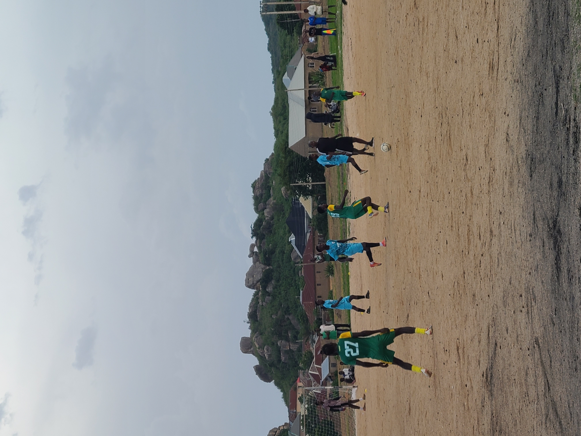 This is a horizontal, eye-level, full shot of a soccer game being played on a dirt field under an overcast sky in Jos, Nigeria. In the foreground, a player in a green and yellow uniform with the number 27 on his back is facing away from the camera, hands at his sides, in a ready stance. To his left, a soccer goal with a white net is visible. Further into the scene, several players in green and yellow and blue uniforms are running and kicking a soccer ball. In the background, a cluster of buildings with red roofs and light-colored walls are nestled at the base of a rocky, green hill. A few people are standing around observing the game. The overall environment is rural, with rough terrain and scattered vegetation.