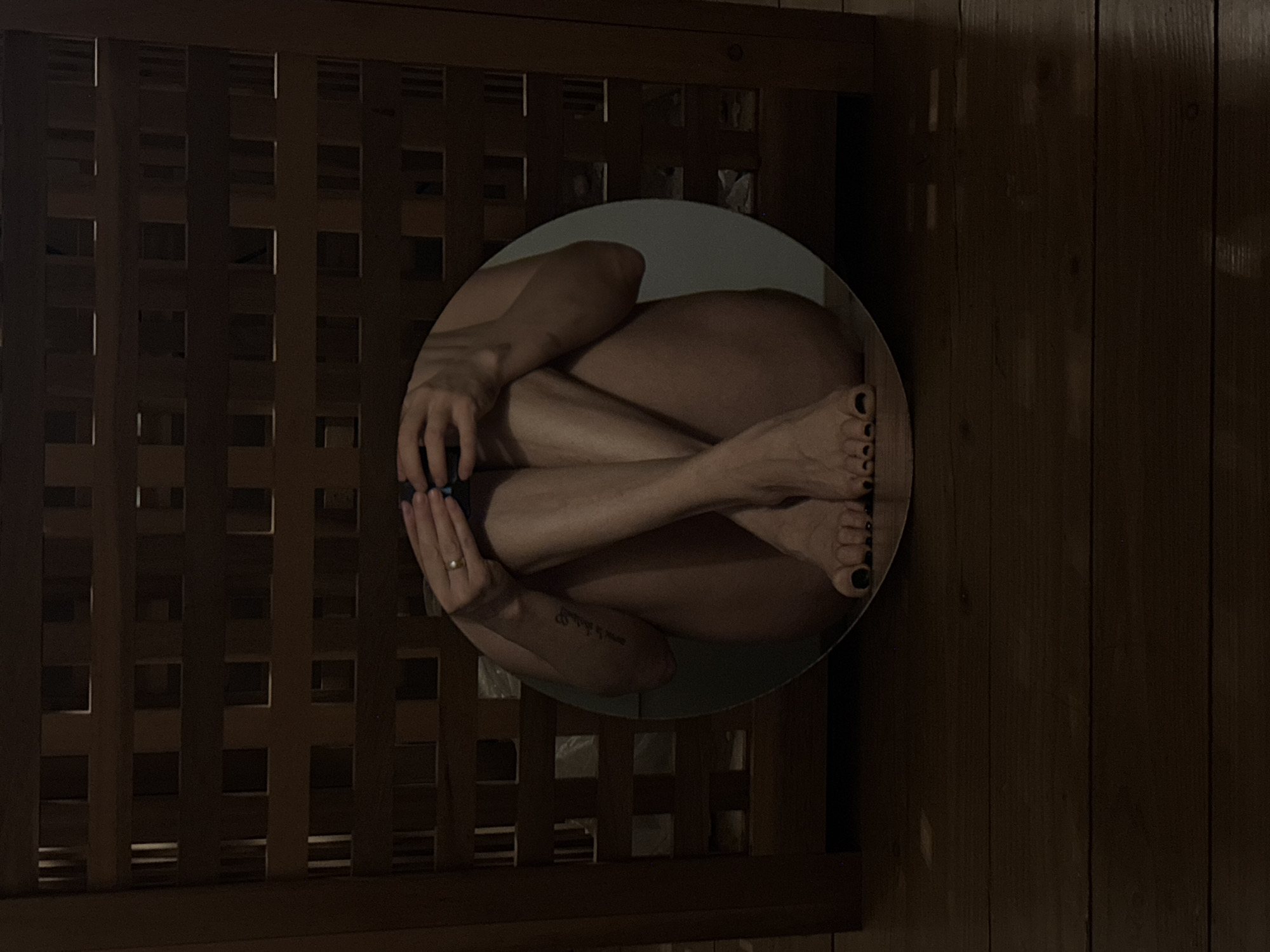 This media file, presented rotated, captures a circular mirror reflecting a person in a curled-up, almost fetal position. The person, appearing nude or wearing minimal clothing, is positioned centrally within the mirror's frame. Their knees are drawn up, and their arms are wrapped around their legs, with their hands holding a dark-colored object, possibly a gaming controller or a small electronic device. A ring is visible on the ring finger of their left hand. Their feet are visible in the reflection, with black nail polish on the toes. Faint, stylized text, likely a tattoo, is discernible on the inner forearm or bicep of the person's left arm.
The background of the image is predominantly dark wood. To the left of the circular mirror, a dark wooden latticework or grid pattern creates a structured texture. To the right, a darker expanse of wooden planks, possibly a wall or floor, is visible.
The lighting is dim and low-key, suggesting an indoor setting, possibly at night or in a dimly lit room, contributing to a private and intimate atmosphere. The scene evokes a sense of introspection or quiet contemplation. The location context for this image is Chaikovsky, Russia. There is no clearly legible text other than the faint tattoo.