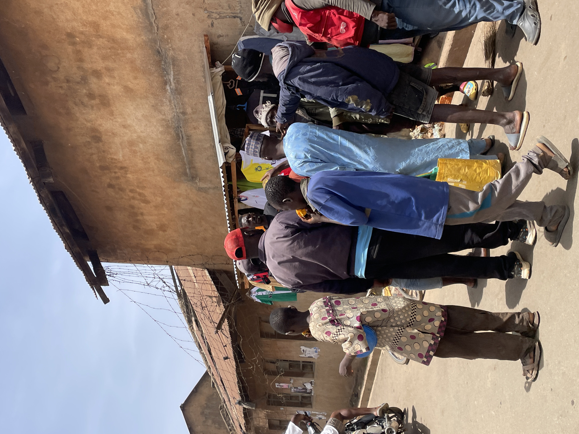 The image captures a lively outdoor street scene in Bukuru, Nigeria, under a bright, clear sky. Numerous people, mostly men, are gathered along a dirt or concrete path, appearing to interact with stalls or shops that line the street.
The setting is characterized by structures made of light brown, possibly mud brick or concrete, material, topped with corrugated iron roofs. Barbed wire is visible along the top of some walls, separating properties. The strong sunlight casts distinct shadows, suggesting the time of day is midday or early afternoon.
Individuals are dressed in a variety of clothing, combining traditional garments with casual modern wear. One man wears a light blue, flowing traditional outfit, while others are in hoodies, jackets, t-shirts, and trousers. Footwear ranges from sandals and slippers to sneakers. Many people are seen standing, some looking downwards, perhaps at items displayed on low tables or within the stalls. Visible goods include various clothing items hanging in the background, hinting at textile or general merchandise vendors.
Notable details include:
- A yellow t-shirt worn by one individual prominently displays the letters "IC".
- Another white t-shirt shows a partially visible "PUMA" logo.
- A small yellow bag or package rests on the ground near one person's feet.
- The front wheel and handle of a white motorcycle or scooter are partially visible in the foreground on the right.
The overall scene depicts a busy, everyday marketplace or street, filled with daily activity and commerce.