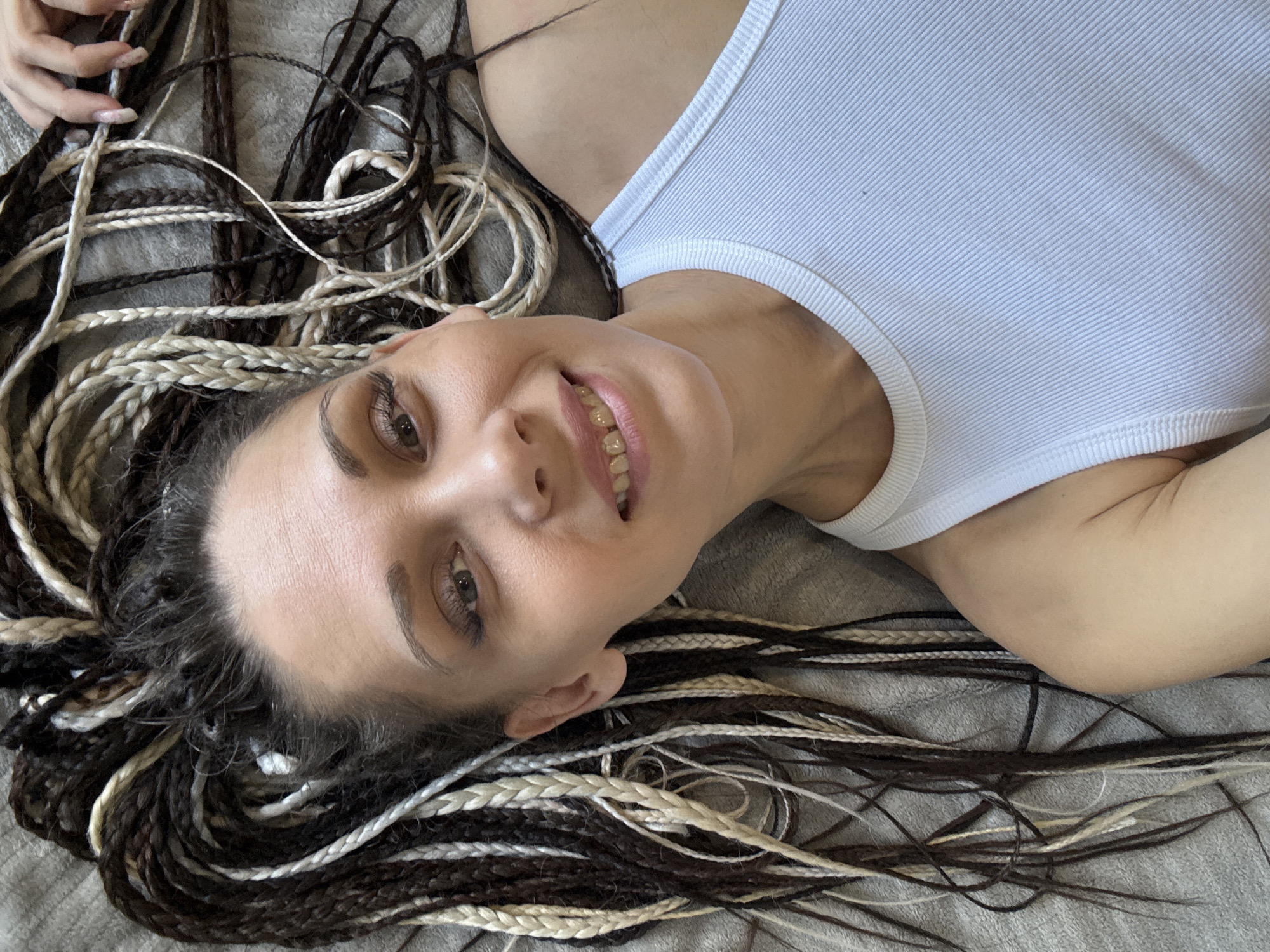 A woman with light skin, light brown eyes, and dark eyebrows and eyelashes is lying on a gray surface. She is wearing a white tank top. Her hair is styled in multiple braids of varying colors, including black, brown, and blonde, spread out around her. She is smiling, revealing her teeth. The location is unknown.