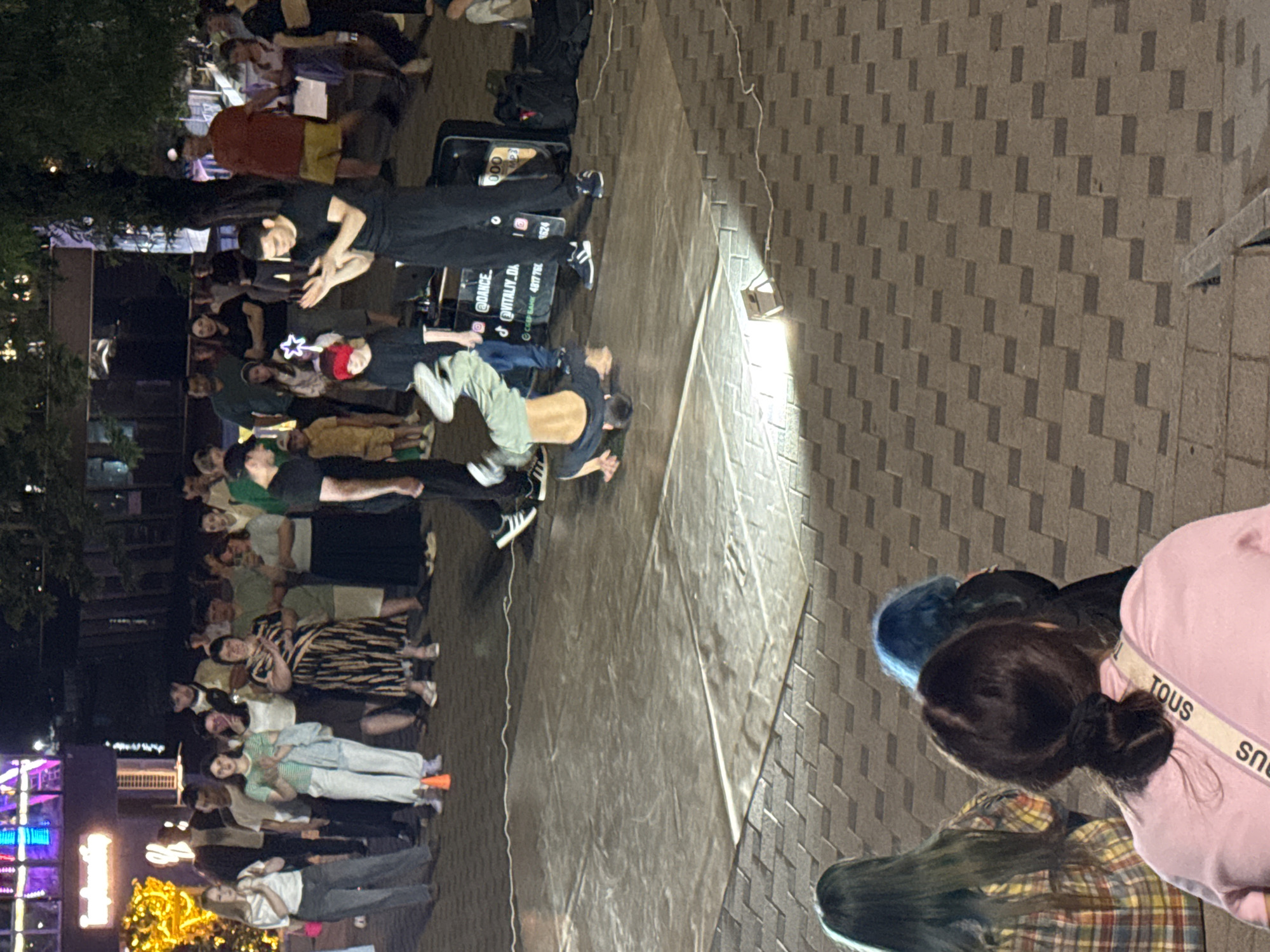 In an outdoor urban setting at night, a group of people are gathered to watch a breakdancer perform on a tarp laid out on a tiled plaza. The breakdancer is in mid-air, performing a handstand with one leg extended. He is wearing a black t-shirt and green pants. Two men in black are standing near the tarp, one with his hands clasped as if applauding and the other in the process of clapping. Behind them, a crowd of spectators, including men, women, and children, observe the performance. Some of the spectators are dressed casually, while others are in more formal attire. In the background, there are buildings with illuminated signs and trees. The plaza is lit by overhead lights and a portable spotlight illuminating the performance area. In the foreground, three people with distinctive hair colors, green, blue, and dark brown, are seated facing away from the camera, also watching the performance. One of them is wearing a pink shirt with a crossbody bag that has "TOUS" written on it.