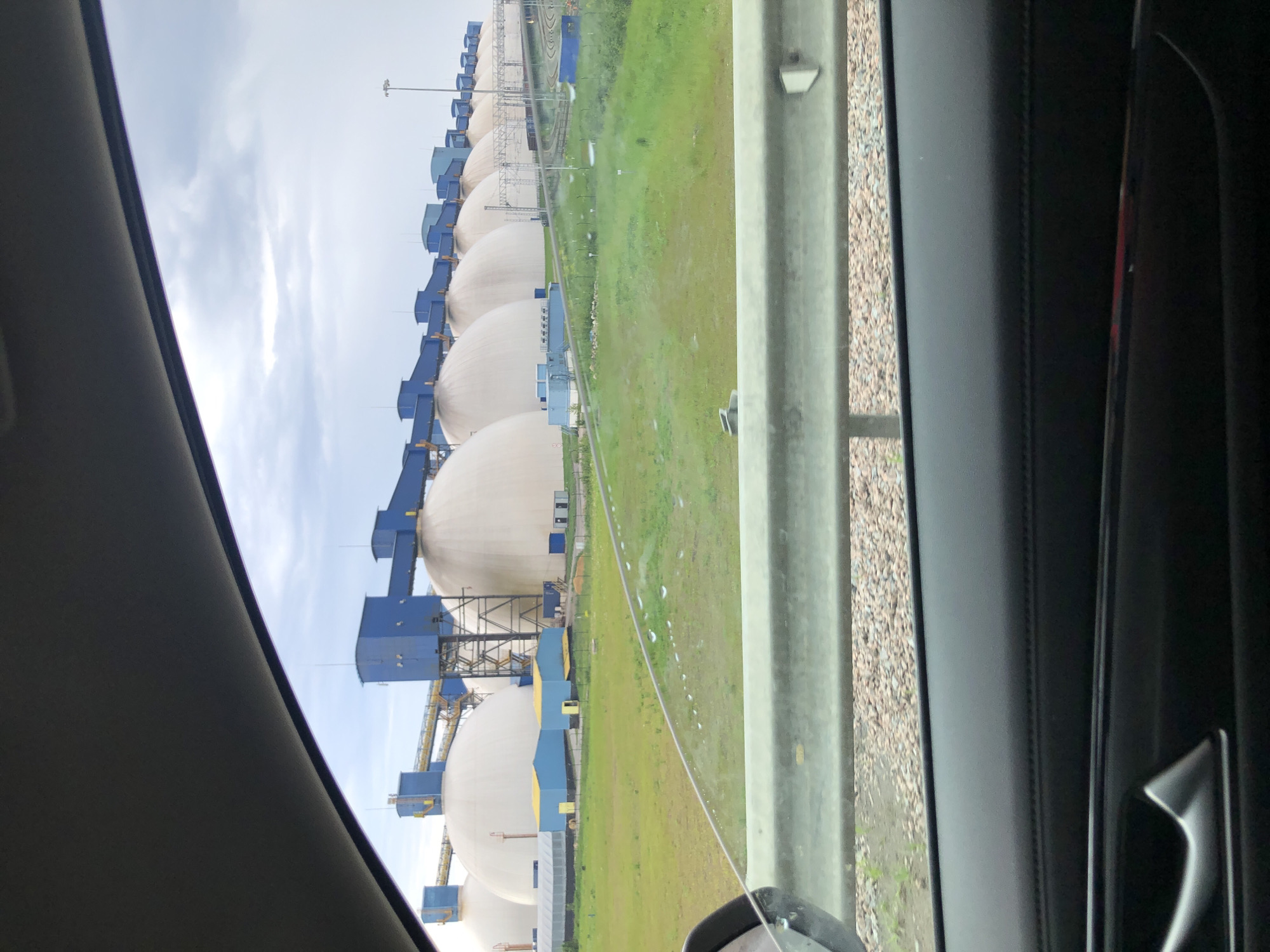 The image displays an industrial scene, likely viewed from inside a vehicle. The prominent subjects are a long row of large, light-colored spherical storage tanks, resembling those used for liquefied natural gas (LNG) or other gases, extending into the distance. Each spherical tank is connected to or accompanied by smaller, blue-colored industrial structures, which appear to house operational equipment, pipework, and support frames. Some of these blue structures feature yellow detailing.
In the mid-ground, a wide, flat expanse of green grass or field separates the viewer from the tanks. There are some lighter, possibly drier or dirt patches within the grass. A grey concrete barrier or guardrail is visible in the foreground, separating the immediate roadside from the field. Beyond this barrier, a gravel-covered area indicates a roadside or shoulder.
The photograph is taken from within a dark-colored vehicle, with parts of the interior, including the window frame and a car door panel with a handle, visible on the left and right sides of the frame. The sky is overcast with light grey clouds. The scene conveys an industrial setting, consistent with the port and energy infrastructure found in Ust-Luga, Russia. No people or discernible text are visible in the image. The time of day is daytime.