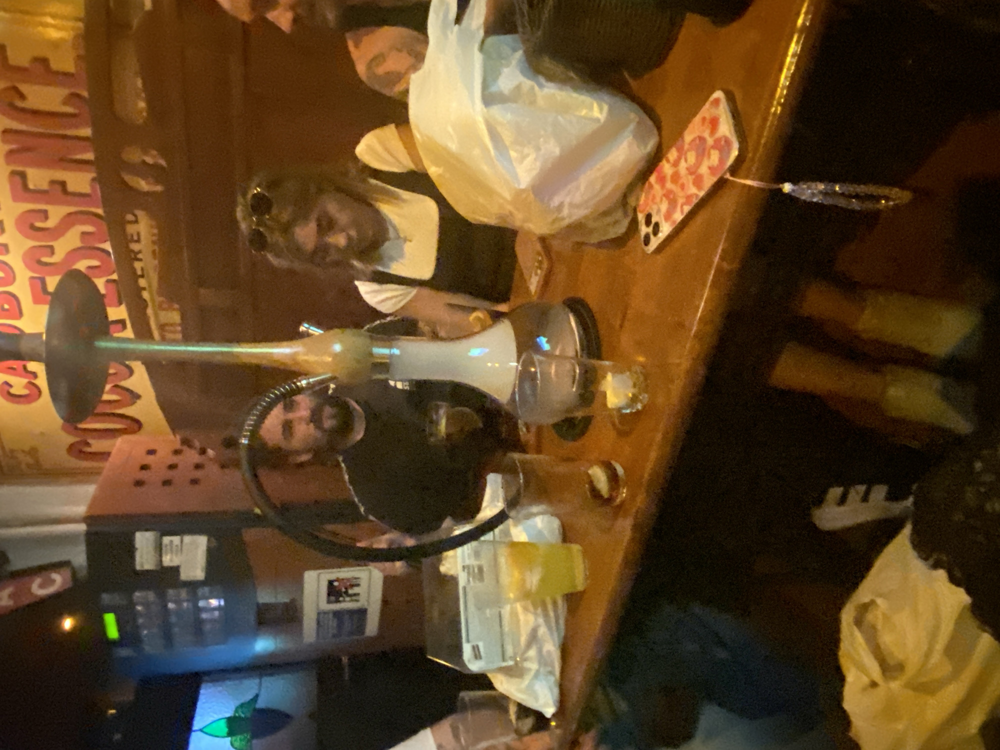 The image depicts a casual bar scene. A hookah pipe is prominently displayed on a wooden bar counter, with two people in the background. A woman is wearing sunglasses and a white top with a black apron, and a man with a beard is also visible. A partially filled glass is nearby, along with a pink cell phone. The bar is decorated with a large sign that reads "COCO ESSENCE."