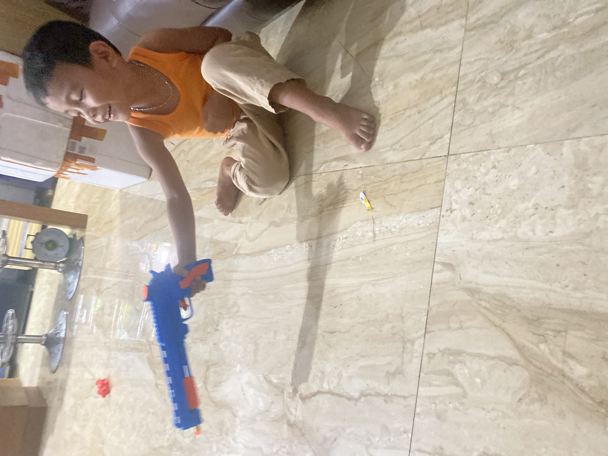 A young boy in an orange tank top and tan pants sits on a marble-tiled floor in a room in Winangun Atas, Indonesia. He holds a blue toy gun with orange accents in his right hand, aiming it forward. The boy is looking to his right and smiling. A small red object is on the floor to his left, and a yellow object is on the floor in front of him. In the background, there are two bar stools with chrome bases and a stack of white boxes with orange trim. A brown leather couch is partially visible on the right side of the image.
