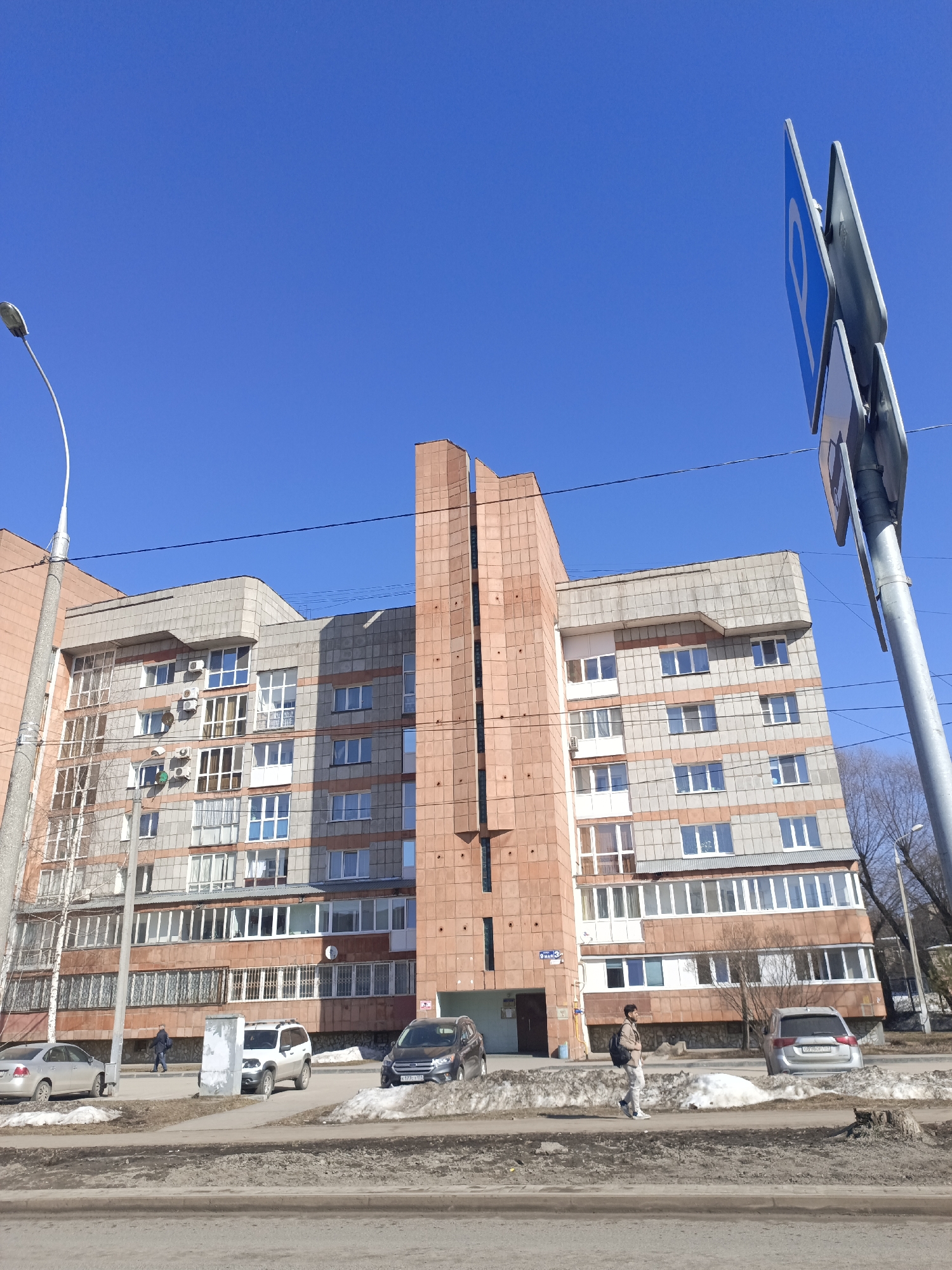 The image depicts an urban scene in Perm, Russia. A multi-story residential building dominates the frame, featuring a mix of gray and reddish-brown brickwork. The building has several windows, some with balconies. A prominent vertical element, clad in reddish-brown tiles, runs up the center of the building's facade.
Several vehicles are parked along the street in front of the building: a silver sedan, a light-colored SUV, and a dark-colored SUV. A pedestrian walks along the sidewalk. A street sign with a blue "P" symbol on it is visible to the right of the building. A lamppost is on the left side. Patches of snow remain on the ground. The sky is clear and blue.