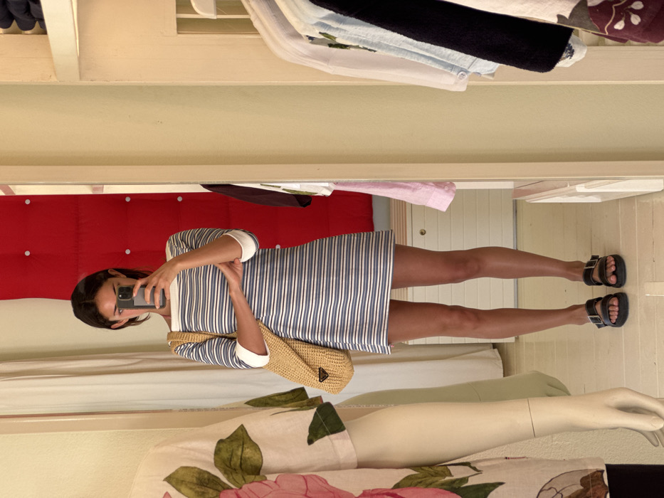 A young adult female is depicted in a full-length mirror, taking a selfie with a dark-colored smartphone that obscures her face. The scene is set inside what appears to be a retail store or fitting room in Marina Maroma, Mexico. She wears a short, boat-neck dress with horizontal blue and white stripes and white cuffs on three-quarter sleeves, paired with dark, two-strap sandals. A light brown woven straw bag with a dark triangular logo is slung over her left shoulder; her left arm is crossed over her right bicep. Reflected behind her is a vibrant red panel adorned with several circular silver studs. To the immediate left of the mirror, a mannequin's torso and arm are partially visible, clad in a light garment featuring a pink floral and green leaf pattern. To the right, a white shelving unit holds various articles of clothing on hangers, including light blue, white, and dark fabrics. The floor beneath her and visible in the reflection is a light, even tone.
