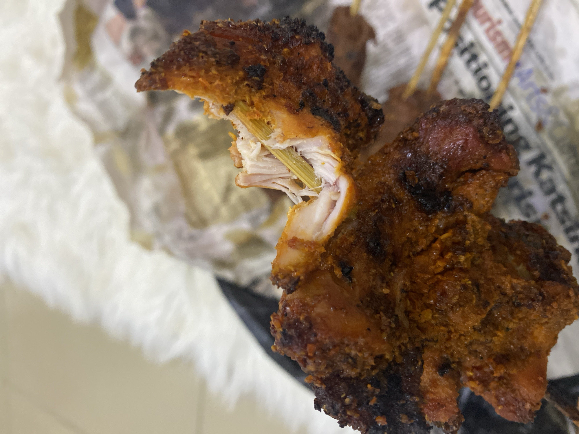 A close-up shot depicts pieces of grilled meat on skewers. The meat has a dark, seasoned exterior. One piece is partly broken apart, revealing pale white meat and the skewer. The background shows crumpled newspaper and a white, possibly fluffy, surface. The image may have been taken indoors. Based on the context, the location could be Jos, Nigeria, but this cannot be confirmed from the image alone.
