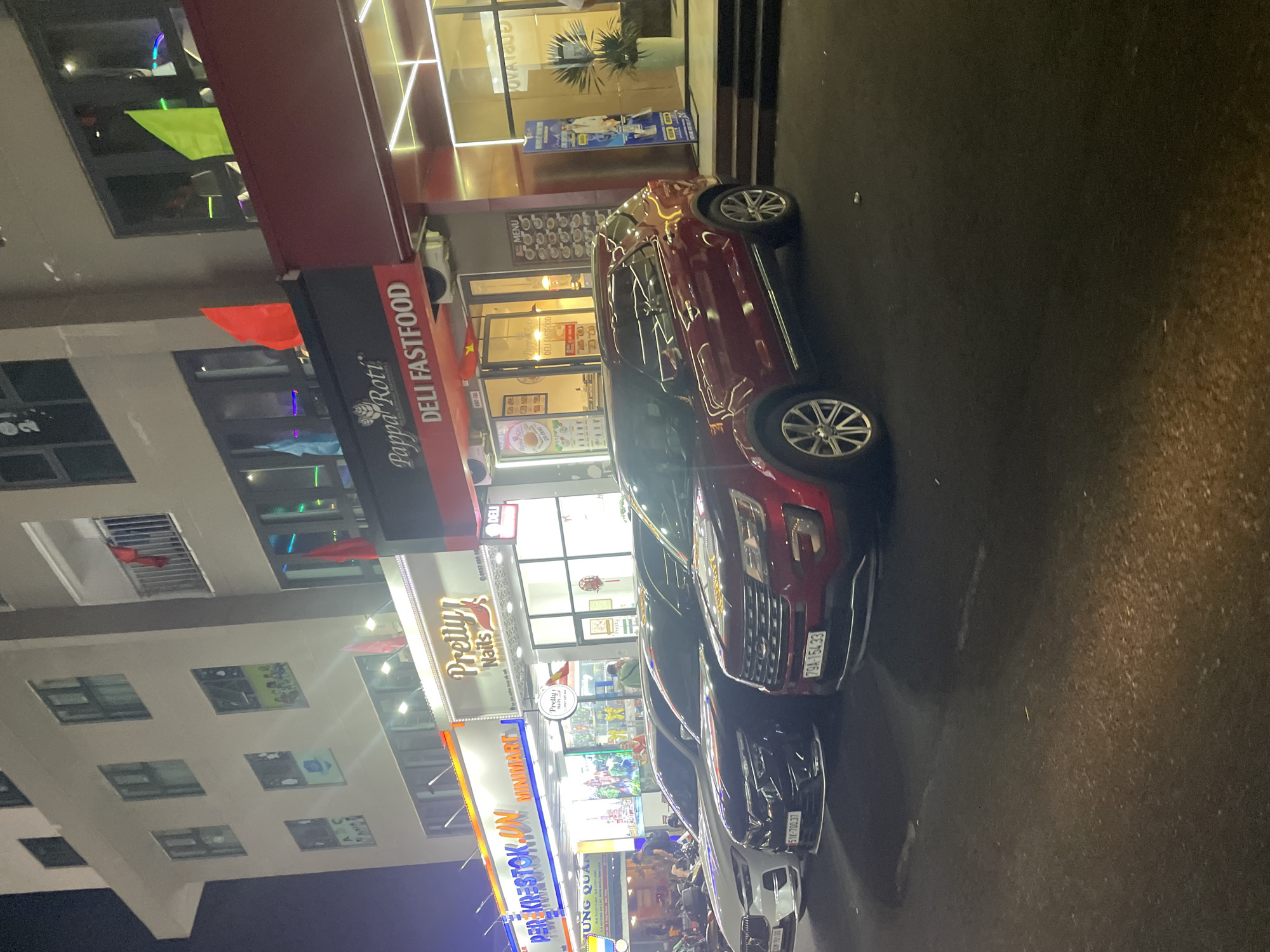 The image captures a street scene in Bắc Nha Trang, Vietnam, at night. The foreground features a line of parked cars, including a red SUV with the license plate "79A-154.33" and a black car with the license plate "51K-700.37". Along the left side of the frame is a multi-story building adorned with signs and storefronts. The signs visible include "Pappa Roti DELI FASTFOOD," "Pretty Nails," and "REFEKRESTOK.UN MINIMART HƯNG QUAN". The storefronts are illuminated, with visible displays and advertisements. Pedestrians and other vehicles appear in the background, contributing to the urban environment.