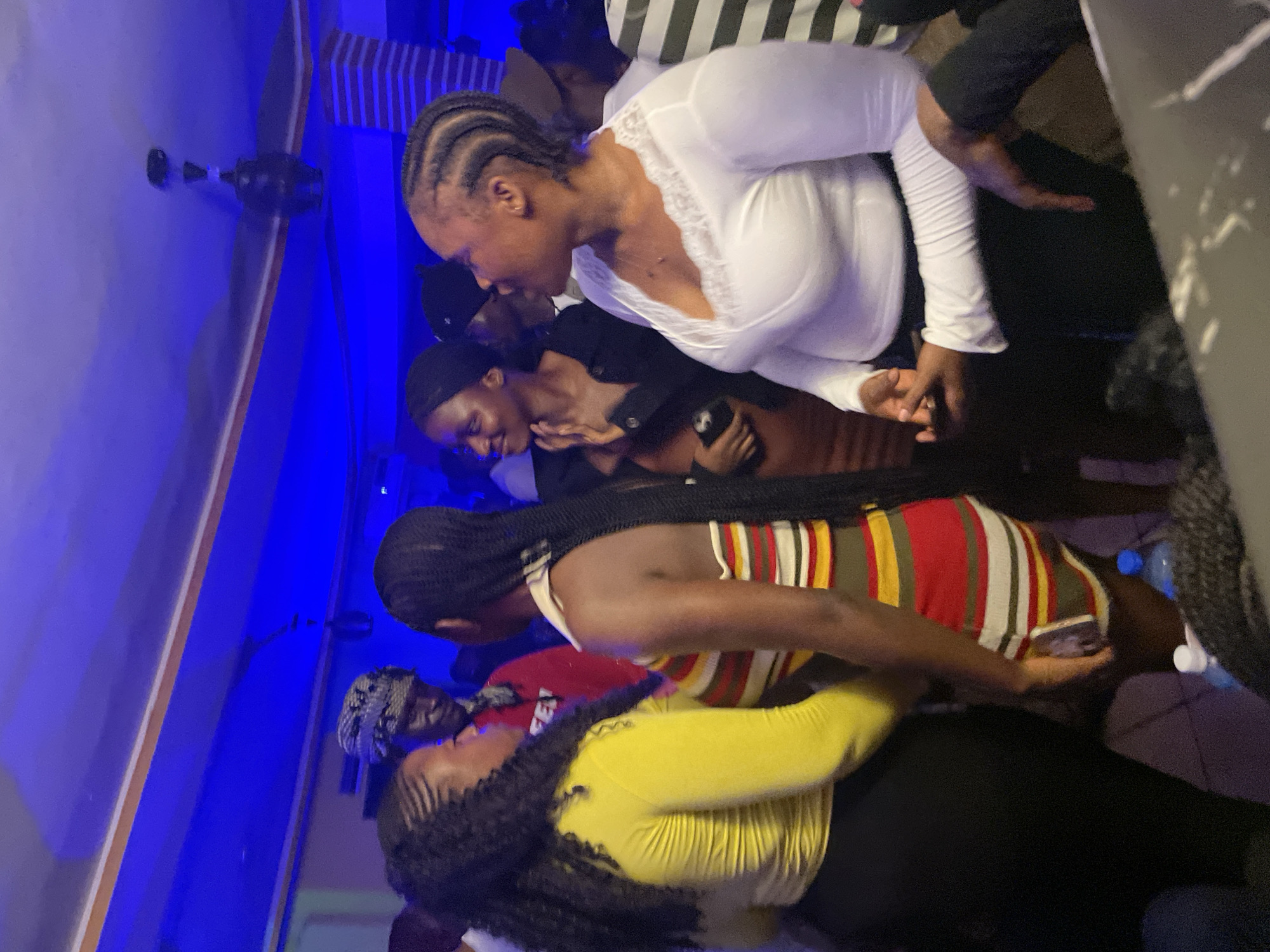 The image depicts a group of individuals in what appears to be a dimly lit, indoor venue, possibly a club or a social gathering. The lighting is predominantly blue, casting a cool tone over the scene.
In the foreground, on the left, a person wearing a bright yellow long-sleeved shirt and a black skirt is facing towards the right, engaged in conversation with another individual. This second person, in the center, is wearing a striped dress with horizontal bands of red, yellow, green, and white. They are also facing left, with their left arm bent and resting on their hip.
To the right of the striped dress wearer, a person in a white long-sleeved top with lace detailing around the neckline is looking to their left, with their hands clasped in front of them. Behind them and further to the right, another person wearing a black and white striped shirt is visible.
In the background, several other individuals are present, some partially obscured. One person in the mid-background is wearing a red hooded jacket and a patterned headwrap. The environment features a ceiling with what appears to be a fan, and brick-like textured pillars. The overall atmosphere suggests a social event. The image does not provide any explicit information about the city of Wumat, Nigeria, or its specific location within it.