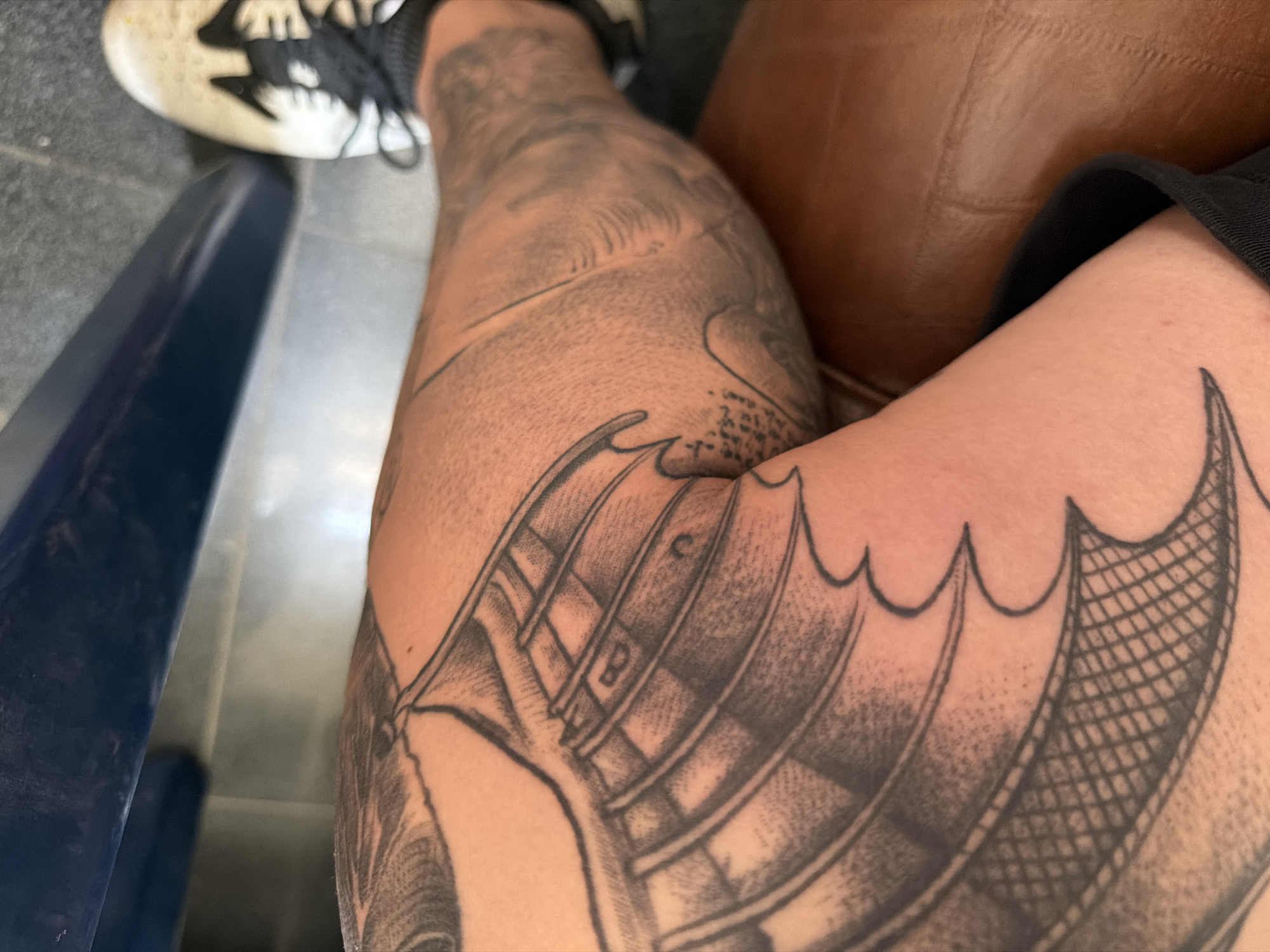 The image shows a close-up view of a person's tattooed leg. The tattoo appears to be a black and grey design, possibly depicting wings or scales with intricate patterns. The leg is resting on a brown, textured surface, possibly leather upholstery. In the background, a white athletic shoe with black laces is visible. The setting suggests an indoor environment, but specific details about the location in Dolno Ablanovo, Bulgaria, are not discernible from the visual information provided.