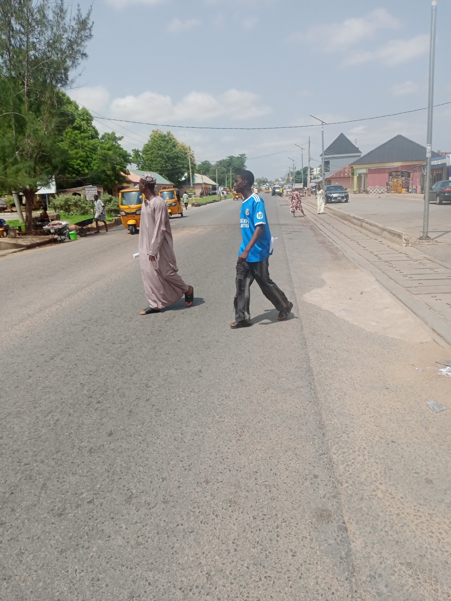 The image depicts a street scene in Jalingo, Nigeria. Two men are crossing the road. The man on the left is wearing a traditional brown robe and a cap, walking towards the right of the frame. The man on the right is wearing a blue t-shirt with white lettering and dark pants, also walking towards the right. In the background, there are several yellow auto-rickshaws, also known as "keke," and other vehicles on the road. Buildings line the right side of the street, some of which appear to be shops. Trees and vegetation are visible on the left side of the road. The sky is partly cloudy. The overall impression is that of a busy, sunny day in a commercial area of the city.