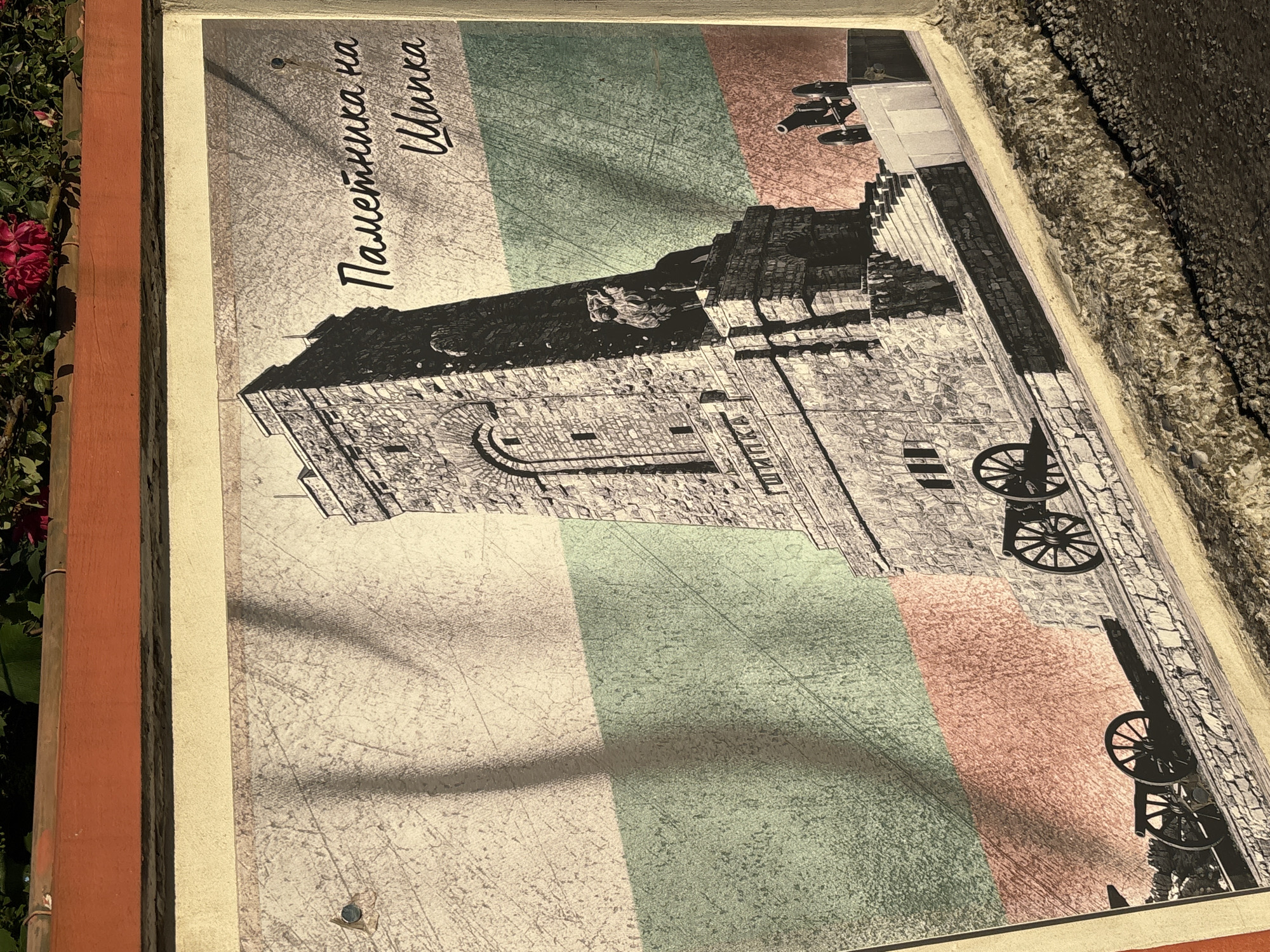 The image displays a monument in the city of Enevo, Bulgaria, against the backdrop of the Bulgarian national flag. The monument, rendered in a sepia tone, is a stone tower with intricate carvings and features a lion statue at its base. Cannons are positioned on either side of the monument, suggesting a historical or military context. The flag's white, green, and red stripes are visible behind the monument, with diagonal shadows cast across the scene. The overall composition is reminiscent of a vintage postcard or historical photograph. The text on the image reads "Паметника на Шипка," which translates to "Monument to Shipka."
