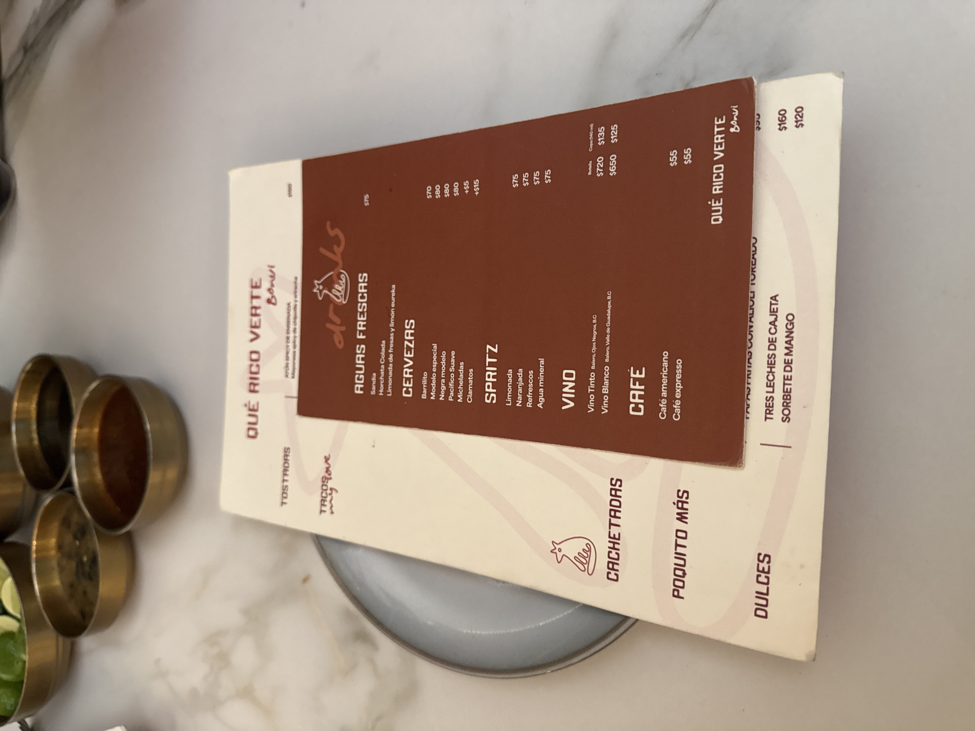 The image shows a restaurant menu from "Qué Rico Verte Bonvi" in Mexico City, Mexico. The menu is placed on top of a plate and next to several small bowls. The menu includes various drinks and food items, such as fresh fruit waters, beers, spritzers, wine, coffee, and desserts. The dessert options include "Tres Leches de Cajeta" and "Sorbete de Mango."