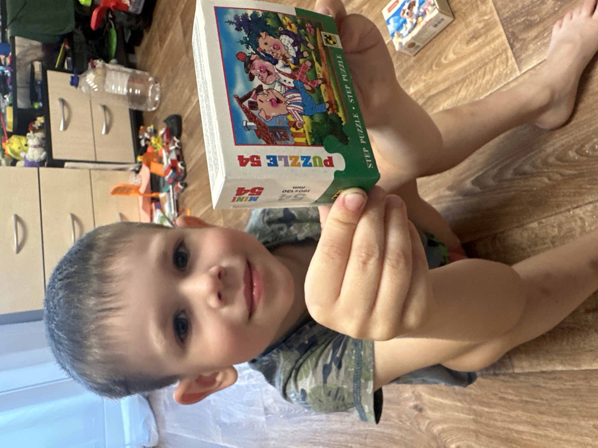 A young boy, appearing to be around 4-6 years old, sits on a wooden floor in what appears to be a home environment. He is wearing a camouflage t-shirt and is holding a small box for a "Puzzle 54" with an illustration of three pigs in overalls, some houses, and trees. The boy's left hand is holding the puzzle box steady while his right hand is gripping it from the side. The background shows a chest of drawers with various toys and a plastic bottle on top. The overall scene suggests a casual, indoor setting. The location in Novoaltaysk, Russia is not discernible from the image.