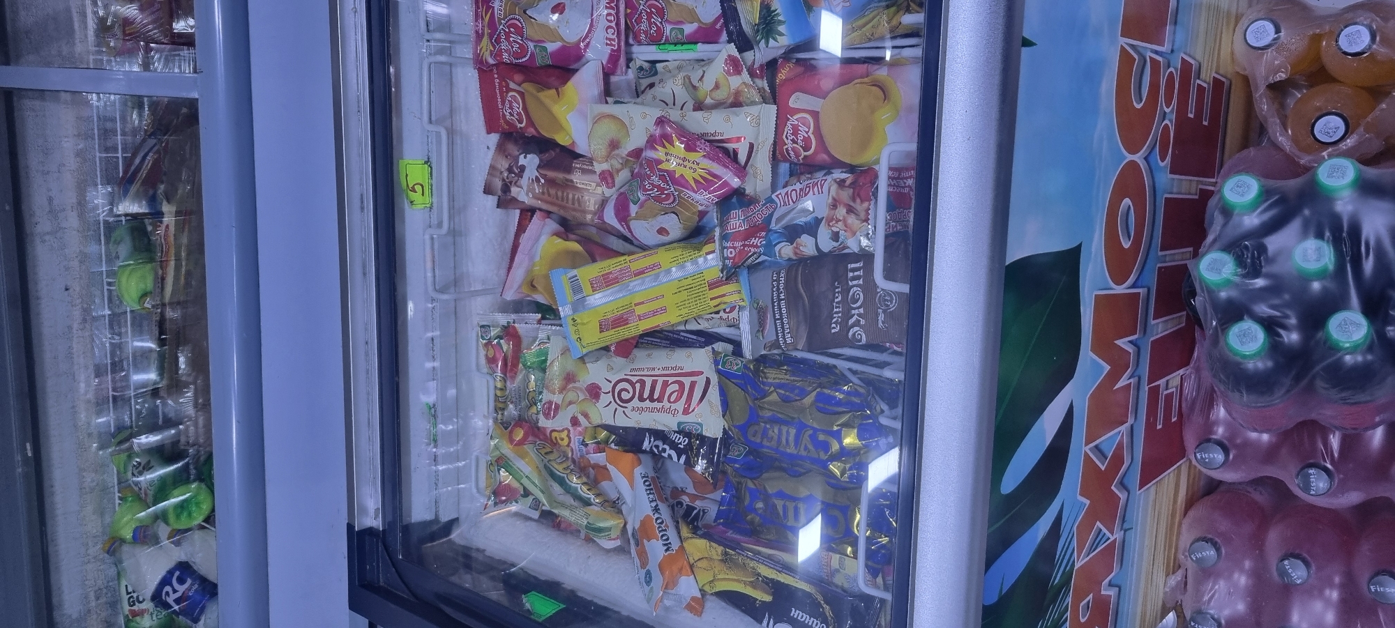The image shows the inside of a commercial freezer displaying a variety of ice cream and frozen treats. The freezer is stocked with individually wrapped ice cream bars, cones, and other frozen desserts in various colorful packaging. Some of the wrappers have text and images, including depictions of fruits like peaches and berries, as well as text in Cyrillic script, suggesting the origin of the products. The lighting inside the freezer is dim, typical of a retail environment, and the glass door of the freezer reflects some of the surroundings. Below the freezer, there are several large plastic bottles filled with liquid, possibly beverages, covered in plastic wrap. The overall scene depicts a retail display of frozen goods in a commercial setting. Based on the provided context, this scene is located in the city of Ismoili Somoni, Tajikistan.