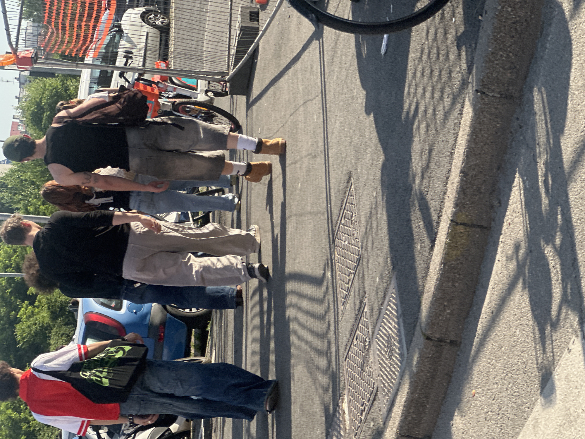 A medium shot reveals four pedestrians on a city sidewalk near a street in Milan, Italy. The pedestrians are captured from the knees up, allowing for detailed observation of their clothing and accessories. The foreground features a close-up of two manhole covers, exhibiting a distinctive diamond pattern. A bicycle tire is partially visible above the group, casting a shadow. The scene is illuminated by the strong natural light.
The foremost pedestrian is dressed in a red and white top and blue jeans, and carries a black bag. Behind, a pedestrian in blue jeans is partially visible. Next is a person wearing a black t-shirt, beige trousers, and black shoes. Finally, a man in a black shirt, grey trousers, tan work boots, and a backpack is depicted. The pedestrians are walking in the same direction.
In the background are cars, fences, and a tree.