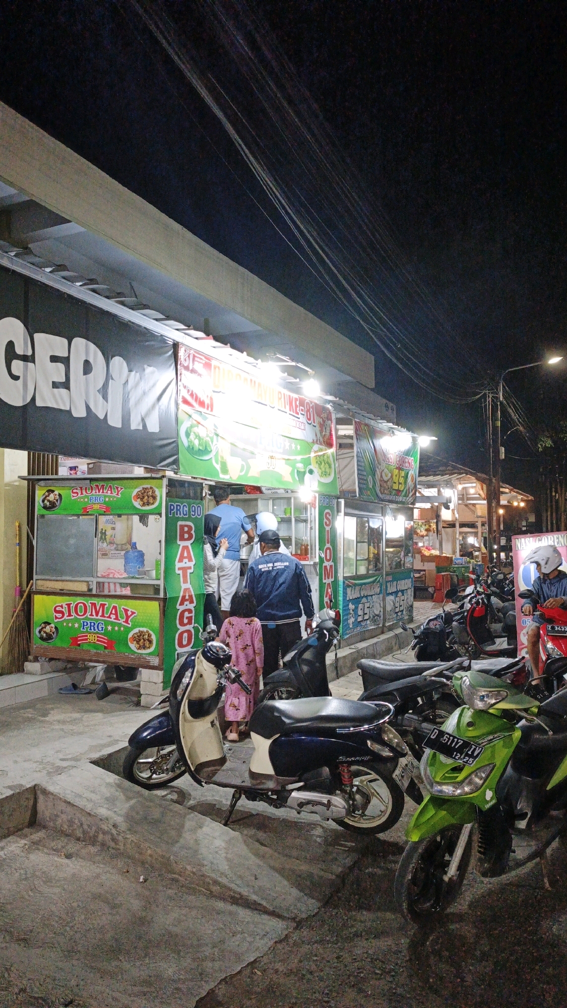 At night, a row of food stalls lines a street in Desa Karangbenda, Indonesia, illuminated by overhead lights. The stalls display banners advertising various food items such as "Siomay" and "Nasi Goreng." Several people are visible, some working behind the counters and others standing nearby. A variety of motorcycles are parked in front of the stalls. The ground is paved, and a concrete embankment runs alongside the stalls. The overall scene depicts a lively street food environment.