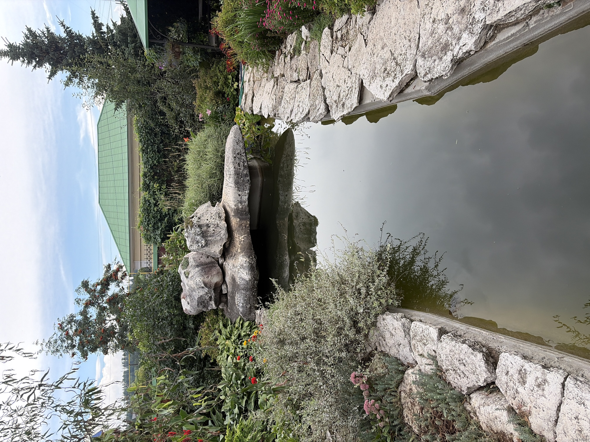 This is a medium-shot, eye-level, vertical, outdoor, daytime, full color photograph of a garden pond in Norja, Russia. The pond is rectangular and lined with large, rough-cut stones. The water is murky and reflects the sky and surrounding foliage. In the center of the pond, a large, irregularly shaped rock formation sits precariously on a narrower stone. Lush green plants and trees surround the pond, with some red and yellow flowers visible on the left side. In the background, a house with a green roof and some electrical wires are visible, along with a tall evergreen tree on the right. The sky is partly cloudy. There are no people or animals visible in the image.