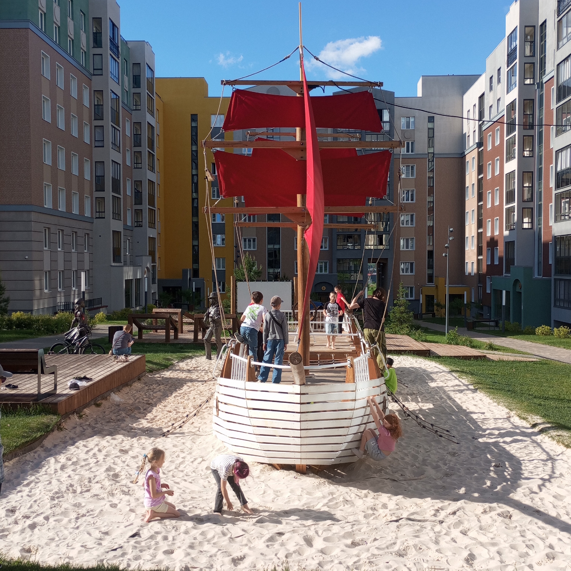 The image displays an outdoor playground set within a modern residential complex, likely in Pervomaysky, Russia. The central feature of the scene is a large, wooden ship-shaped playground, complete with a mast and bright red sails, resembling a pirate or sailing vessel. This ship is situated in a spacious sandy area, creating a beach-like setting.
Several people, including both children and adults, are present. Children are actively playing on and around the ship: some are on the ship's deck, one child with red hair is seen climbing a rope ladder on the side of the hull, and other children are playing in the sand near the ship's bow. Adults are visible supervising on the ship's deck and relaxing on nearby wooden platforms.
The playground area includes wooden benches, platforms, and patches of green grass interspersed with the sand. Two decorative statues are also visible in the midground – one of a person next to a bicycle and another of a standing figure.
The background is dominated by tall, multi-story residential buildings that encircle the playground. These modern buildings feature diverse architectural styles and a palette of colors, including yellow, brown, grey, green, and white facades, indicative of a contemporary urban development.
The scene takes place on a bright, sunny day with a clear blue sky and a few scattered white clouds. Long shadows cast by the ship and buildings suggest it is either mid-morning or late afternoon. The overall atmosphere conveys a sense of lively recreation and community enjoyment. No visible text is legible in the image.