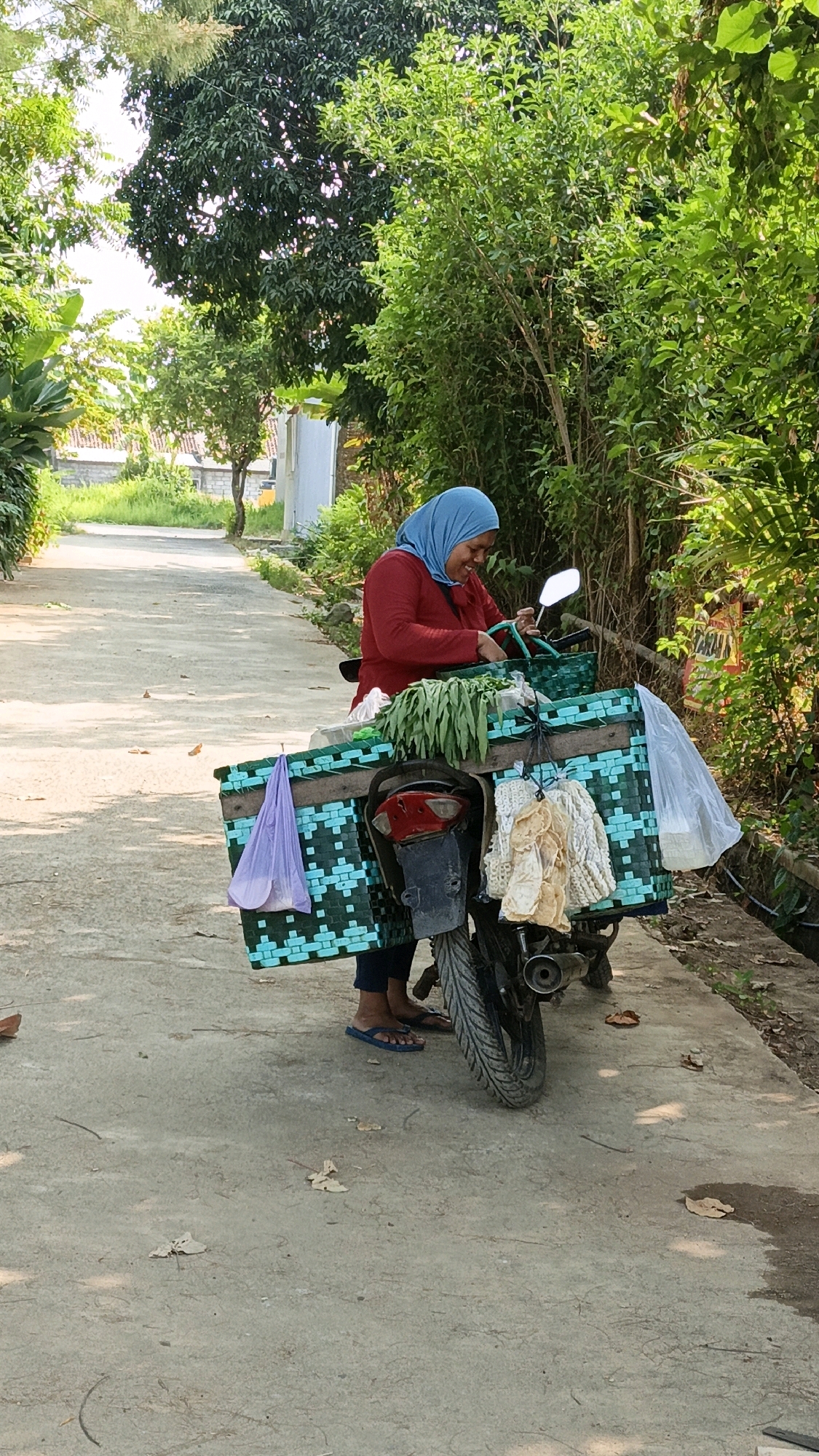 An Indonesian woman wearing a blue hijab and a red top stands beside a motorcycle parked on a paved path in Cikal, Indonesia. The motorcycle is equipped with a large, woven basket filled with green vegetables. A purple plastic bag hangs from the left side of the basket, and several packages of dried goods are attached to the right side. The woman is smiling and appears to be arranging the produce. The environment is lush with green trees and foliage on either side of the path.