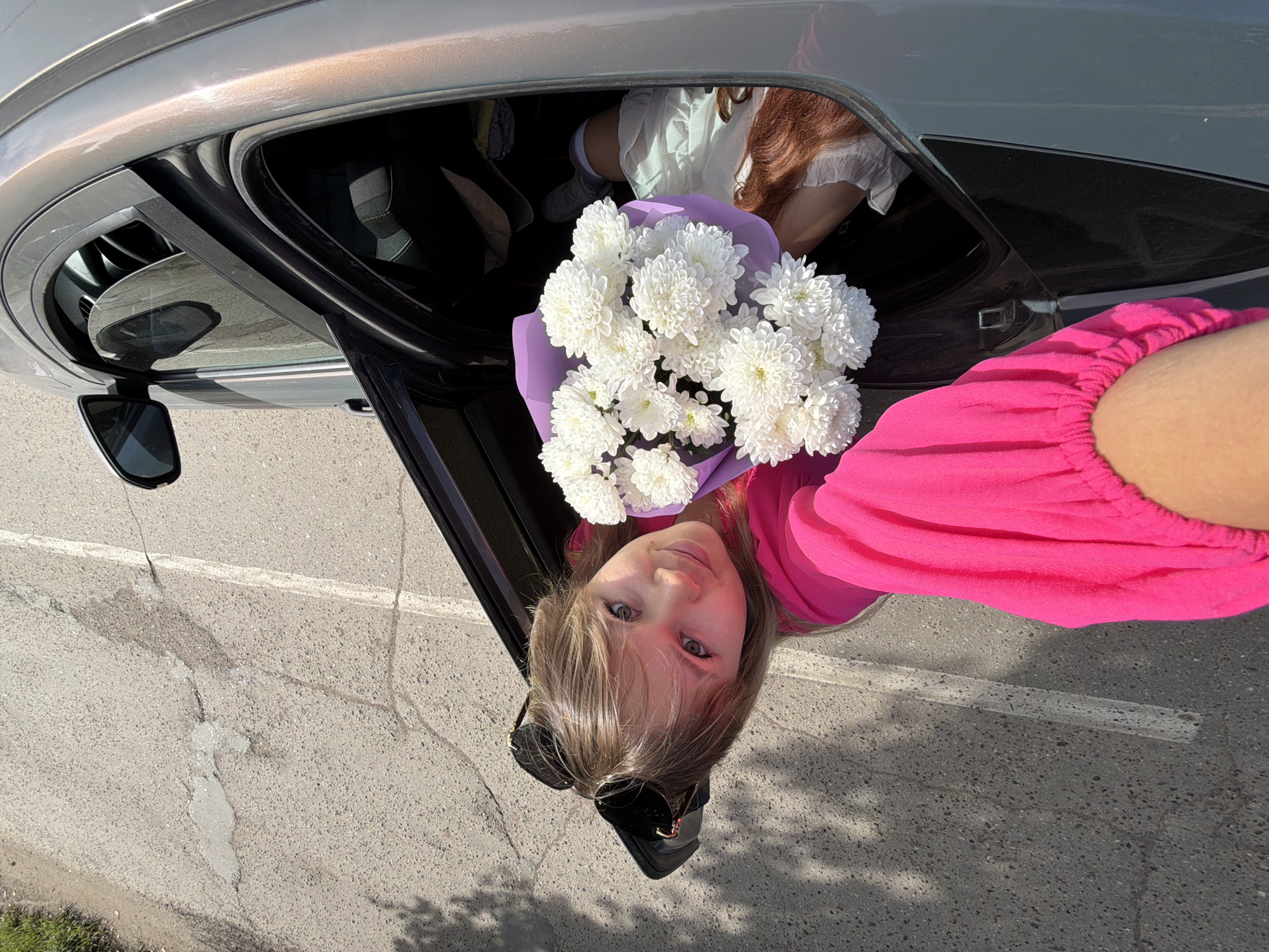 An overhead, close-up, exterior, eye-level shot shows a young blonde woman with sunglasses perched on her head, leaning out of a car door, holding a bouquet of white chrysanthemums. She is wearing a bright pink blouse. Her right arm, partially visible, is extended towards the viewer. Inside the car, partially obscured by the bouquet and the woman, a person with red hair is visible, wearing a white blouse. The car appears to be a gray sedan, and it is parked on a paved road with a white line marking. The road is cracked and shows some signs of wear. The scene takes place outdoors on a sunny day, with shadows cast on the road. The backdrop suggests an urban or suburban environment, consistent with the city of Chaikovsky, Russia, given the context.
