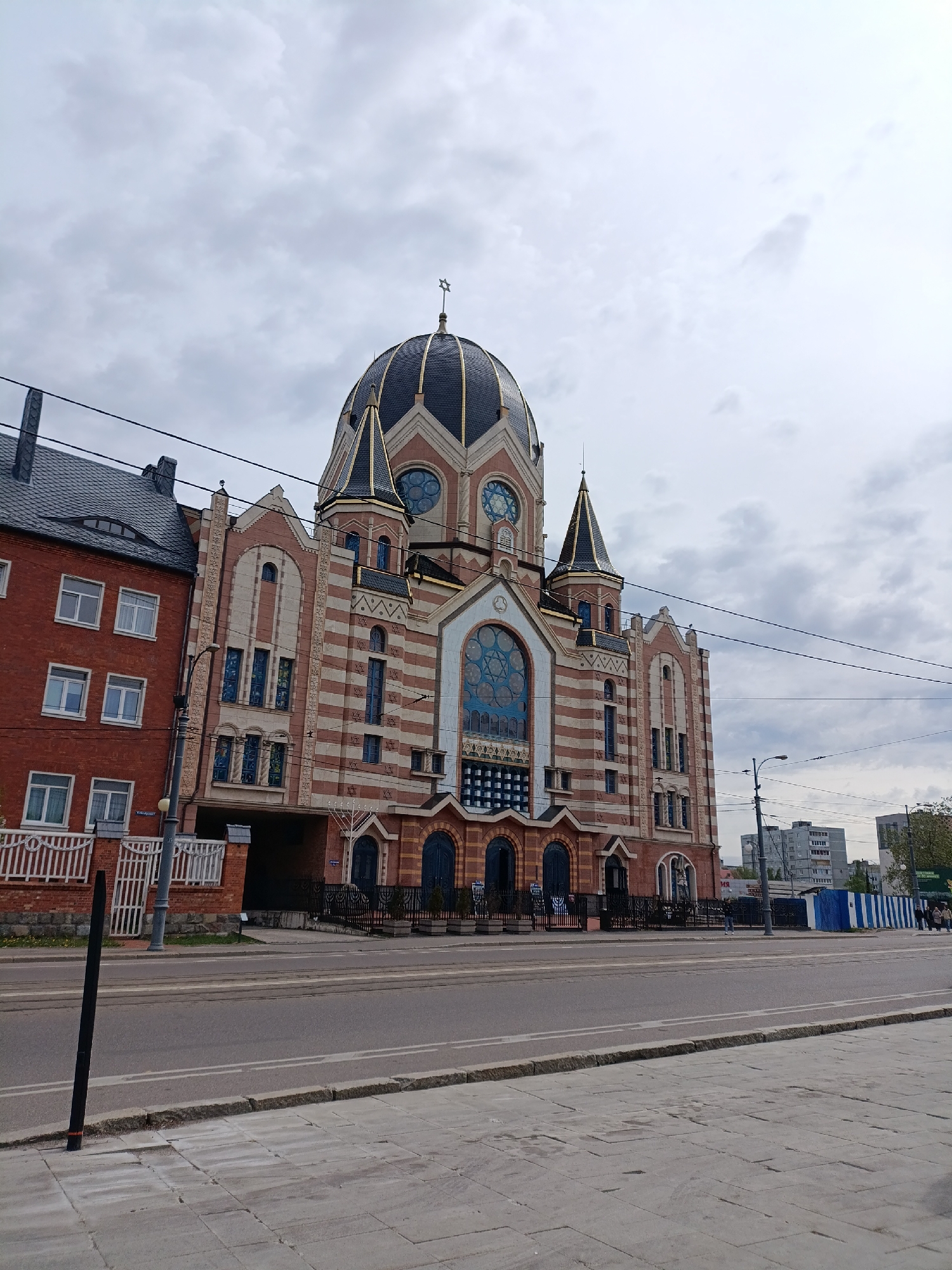 The image captures a street scene in Kaliningrad, Russia, dominated by the New Synagogue, a large brick and stone building with a distinctive dark dome adorned with a Star of David. The synagogue exhibits a striped pattern of alternating red brick and light stone. Adjacent to the synagogue is a smaller brick building with a grey slate roof.
The scene is viewed from across the street. The foreground consists of a sidewalk paved with grey stone blocks and a multi-lane road with tram tracks. Power lines are visible overhead.
In the background, several apartment buildings rise above the synagogue. A blue and white striped structure is on the street behind the synagogue. Pedestrians are walking near the blue and white structure.
The sky is overcast with grey clouds, suggesting a cloudy day.
