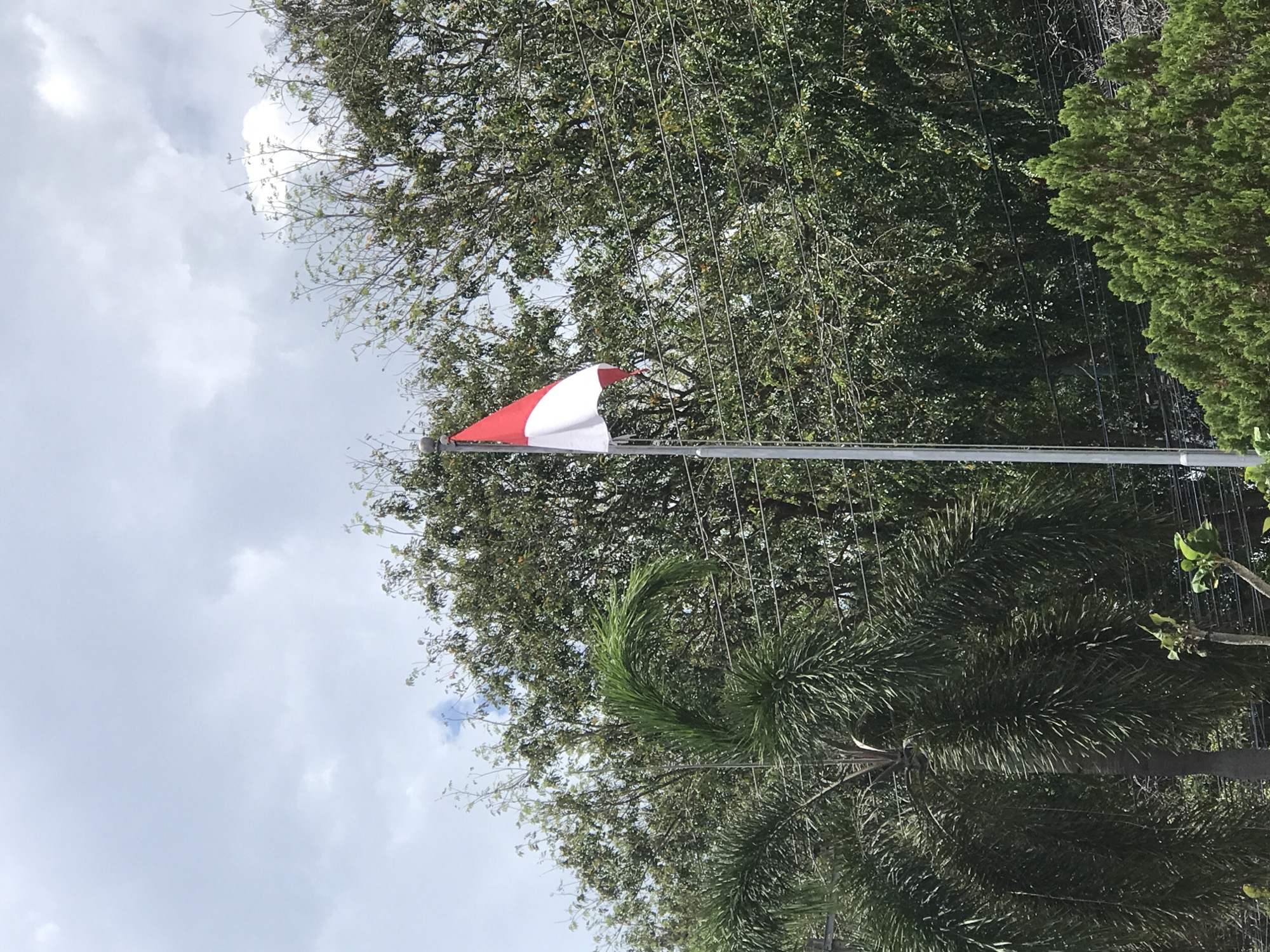The image shows a red and white flag of Indonesia waving on a pole. The sky is visible in the background with some clouds. Trees and other greenery are around the flag. The location is likely Lhokseumawe, Indonesia.