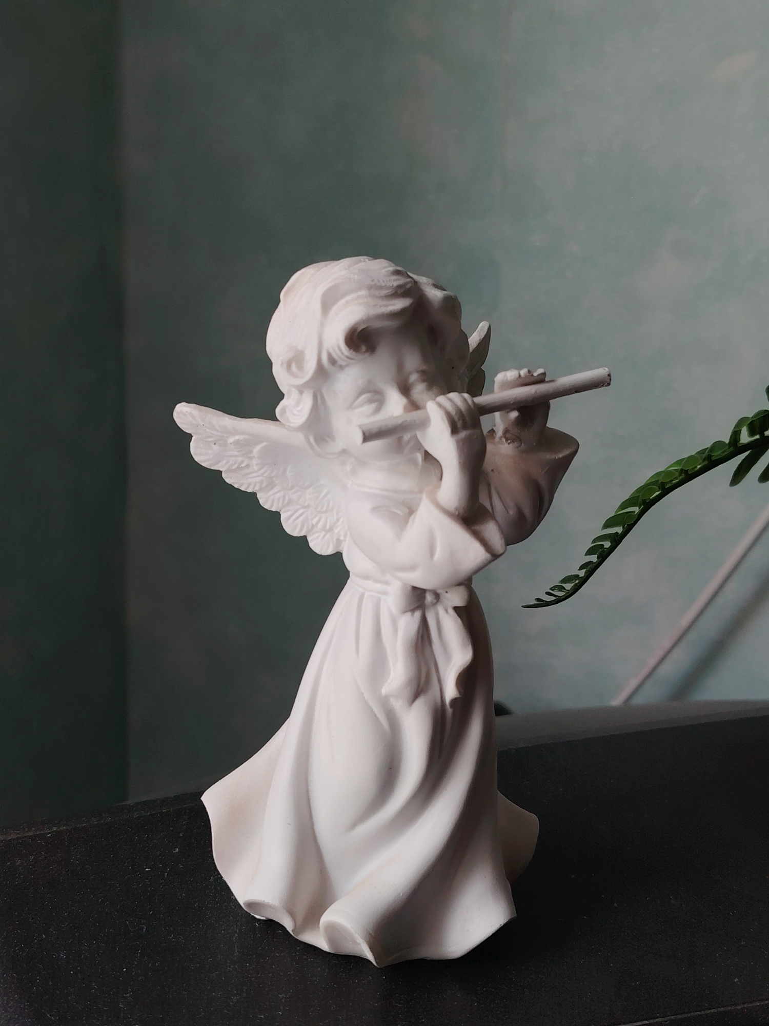 A white angel figurine is placed on a dark, textured surface. The figurine depicts a cherubic figure with wings, wearing a flowing robe and playing a flute. The background is a gradient of muted blue and green. A sprig of greenery with small leaves extends into the frame from the right side of the image, near the figurine. This scene is not in Volnoye, Russia.