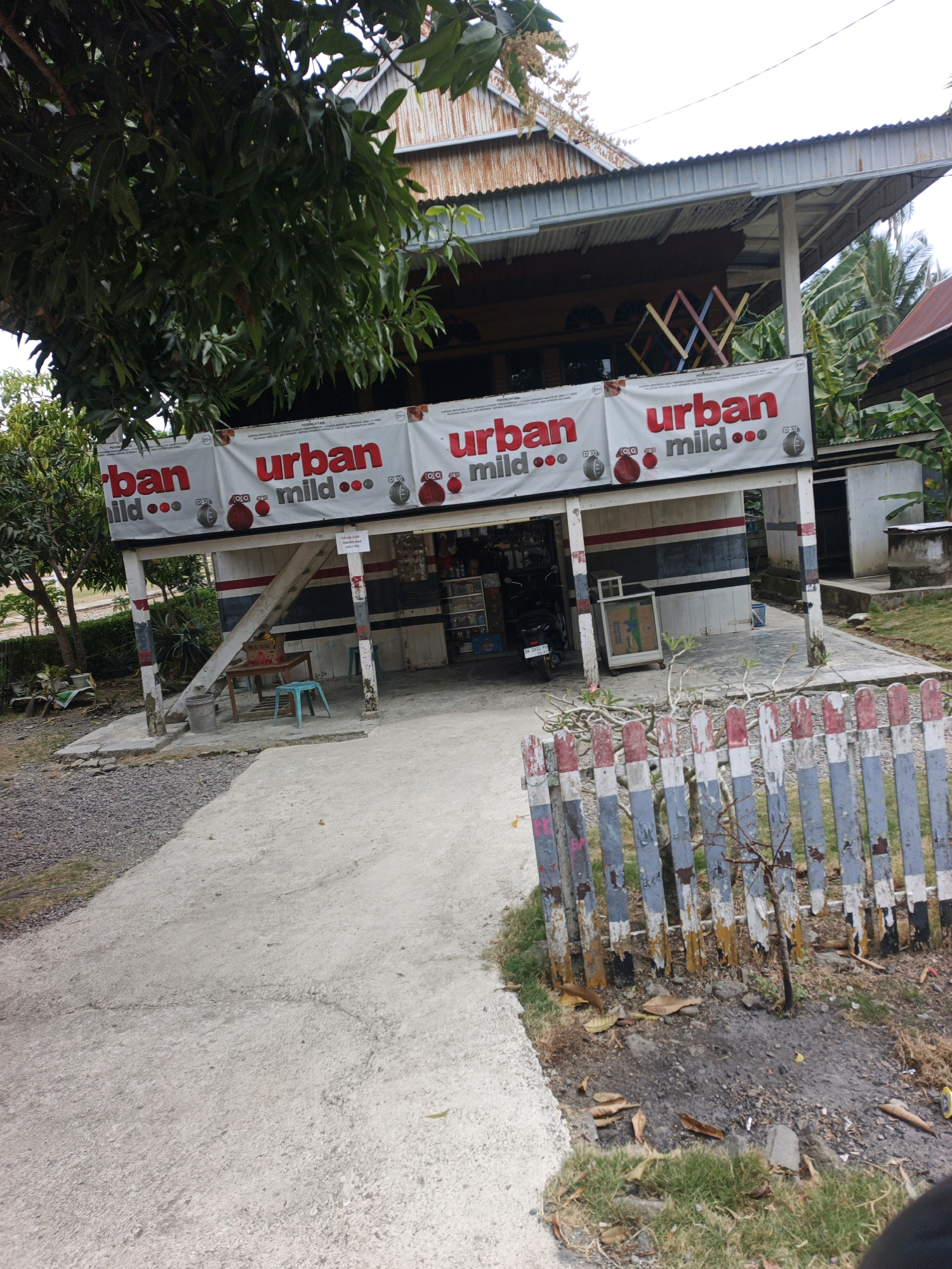 The image depicts the exterior of a roadside convenience store in the city of Laerung, Indonesia. The store has a corrugated metal roof and white walls with red and blue stripes. A banner displaying the brand "urban mild" with images of what appear to be cigarette packs hangs across the front of the store. Several pillars support the roof. A concrete pathway leads to the store, with a small table and a teal stool placed outside. To the left of the entrance, a motorcycle is parked. In front of the store, a dilapidated wooden fence with red and blue painted planks encloses a small area. The ground is covered with a mix of concrete, gravel, and dry grass. Trees and foliage are visible in the background. The scene appears to be an outdoor shot, taken during daylight.