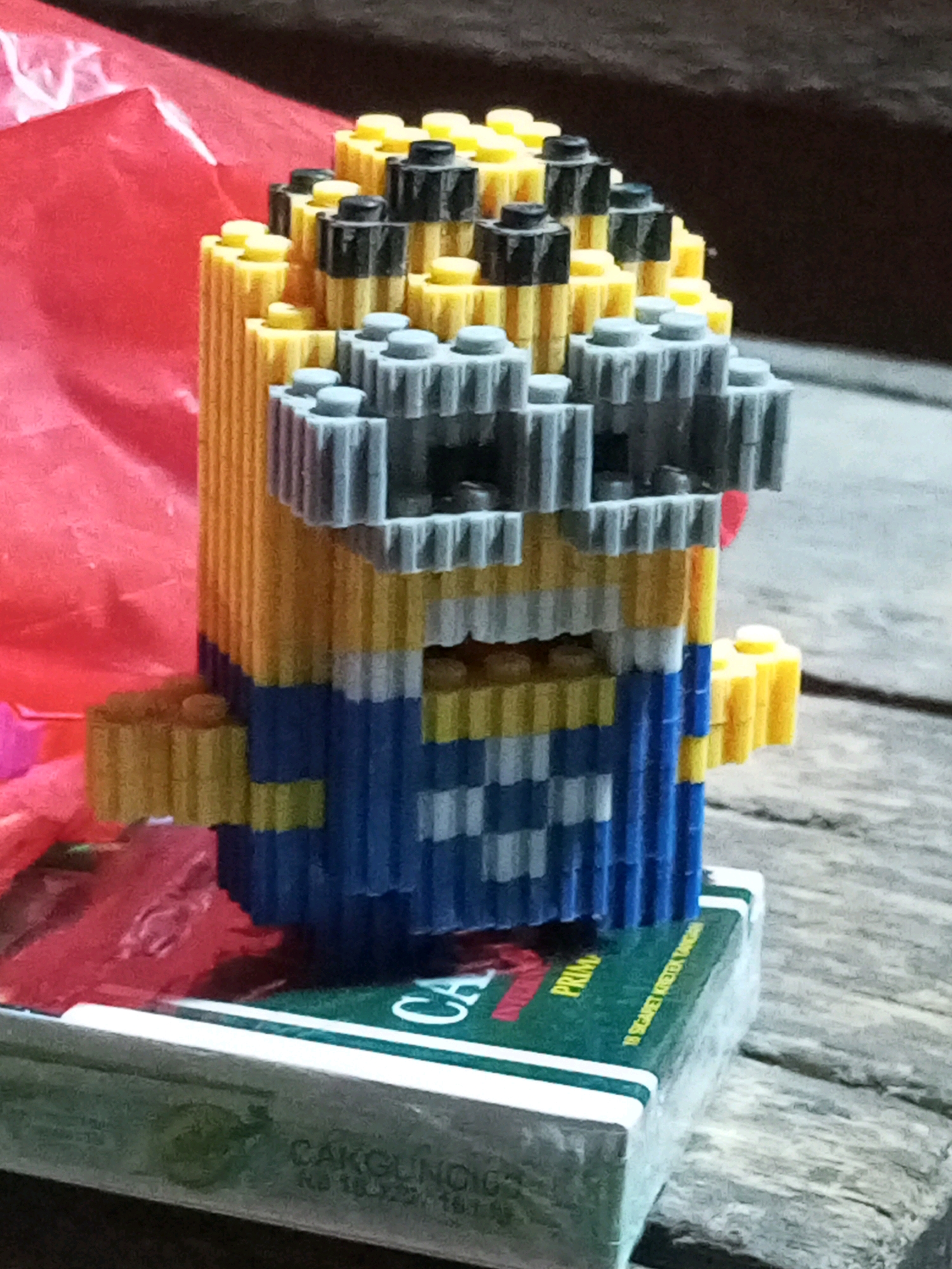 A close-up, eye-level shot shows a minion-like figure constructed from colorful plastic building blocks. The figure is predominantly yellow, with blue sections for its overalls, and grey blocks forming its goggles. The figure is positioned on top of a green and white box that has some text and a red graphic on it. The background is blurred, with a reddish-pink crinkled material to the left and a dark, textured surface to the right, possibly a wooden table. The lighting appears to be artificial and somewhat dim. No people are visible in the image, and no specific location within Keling Panggau, Indonesia, is discernible from the visual cues.