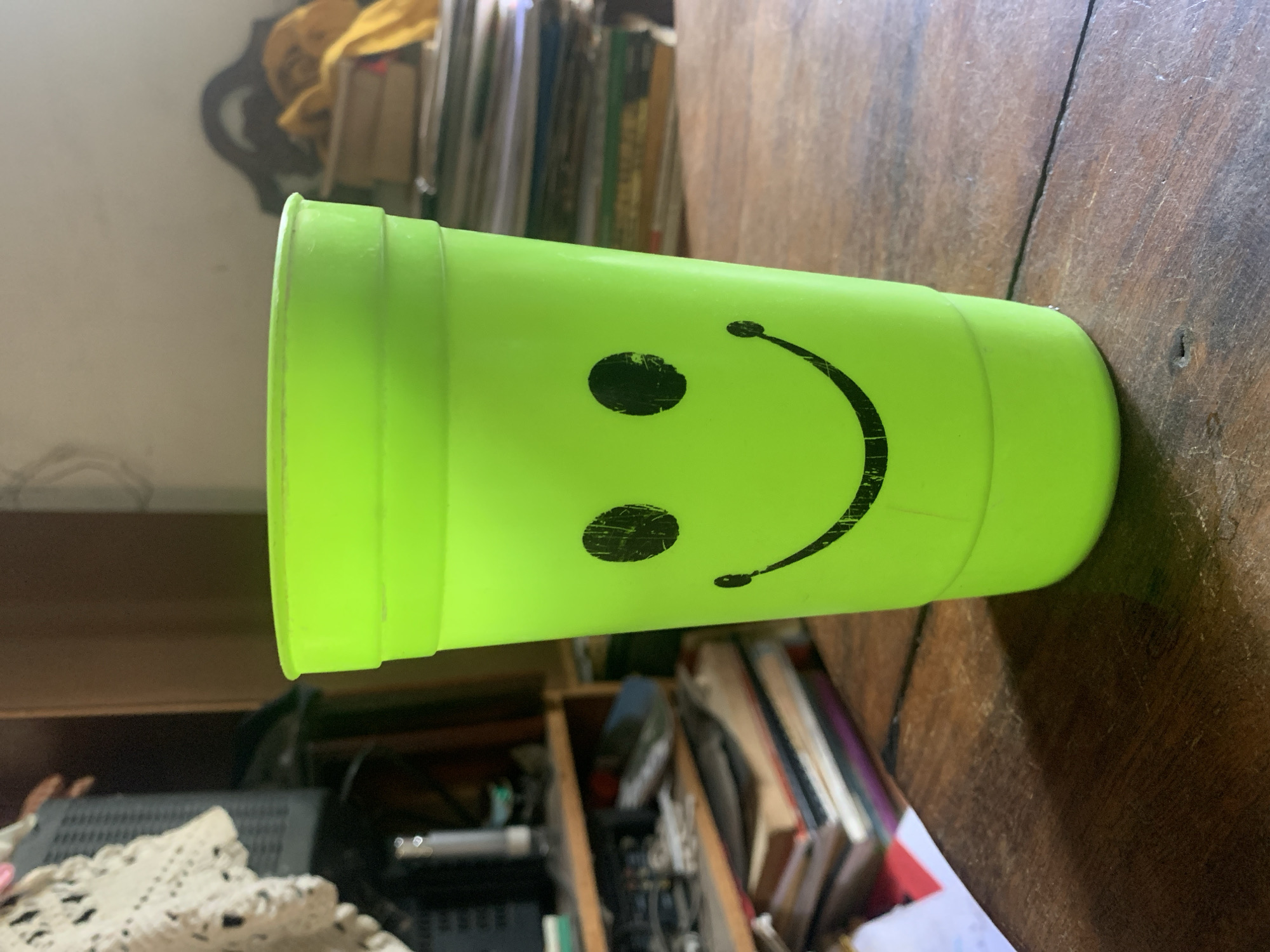 A lime-green plastic cup with a black smiley face is lying horizontally on a dark, wooden surface. The cup appears to be slightly worn. Behind the cup, there is a cluttered shelf filled with books and other items. Further in the background, there are more objects stacked on top of each other. The location is not determinable from the image.