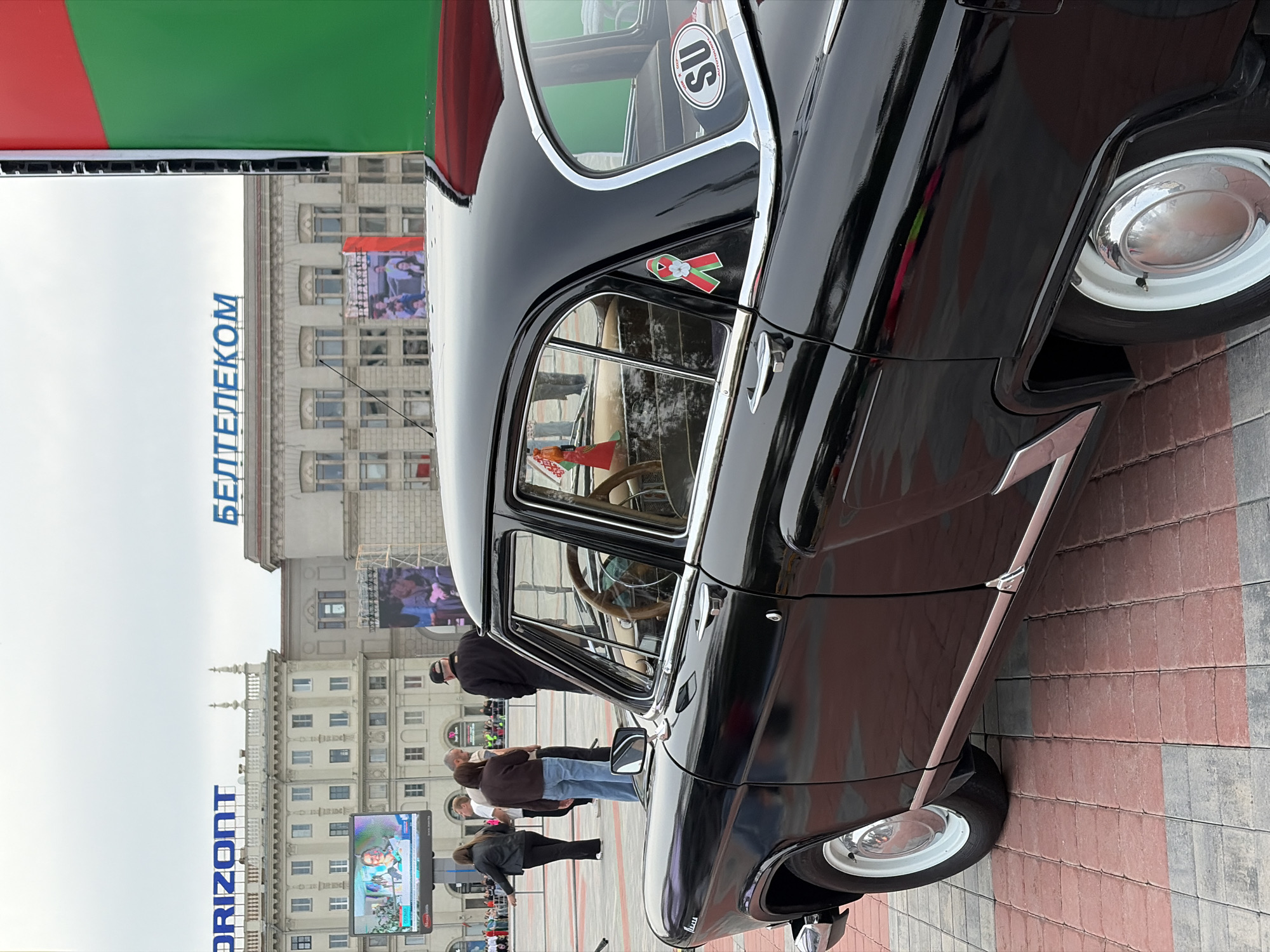 In Minsk, Belarus, on a cloudy day, a vintage black car is parked on a red and gray brick pavement. The car is positioned at an angle, showcasing its side and part of its front. The car features chrome trim, white-rimmed wheels with hubcaps, and a wooden steering wheel visible through the window. A ribbon with green, red, and white colors is affixed to the car's body near the rear window, along with a sticker that reads "SU." In the background, buildings with "ОRIZONT" and "БЕЛТЕЛЕКОМ" signs are visible. People are gathered in the background, some standing and conversing, others walking. A large screen displaying an image is also present. The overall scene depicts an outdoor event or gathering.