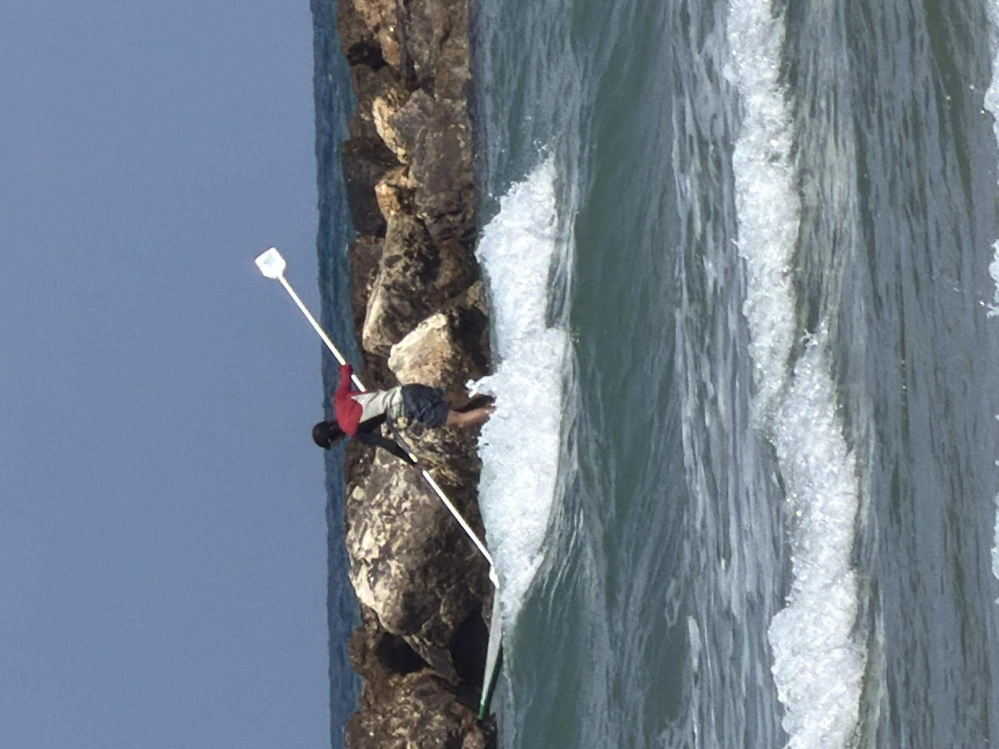 The image depicts a person on a rocky breakwater adjacent to a turbulent sea, seemingly fishing with a long rod, against the backdrop of a clear sky. The waves crash against the barrier. The location is plausibly Tel Aviv, Israel, given the coastal environment and architectural characteristics.