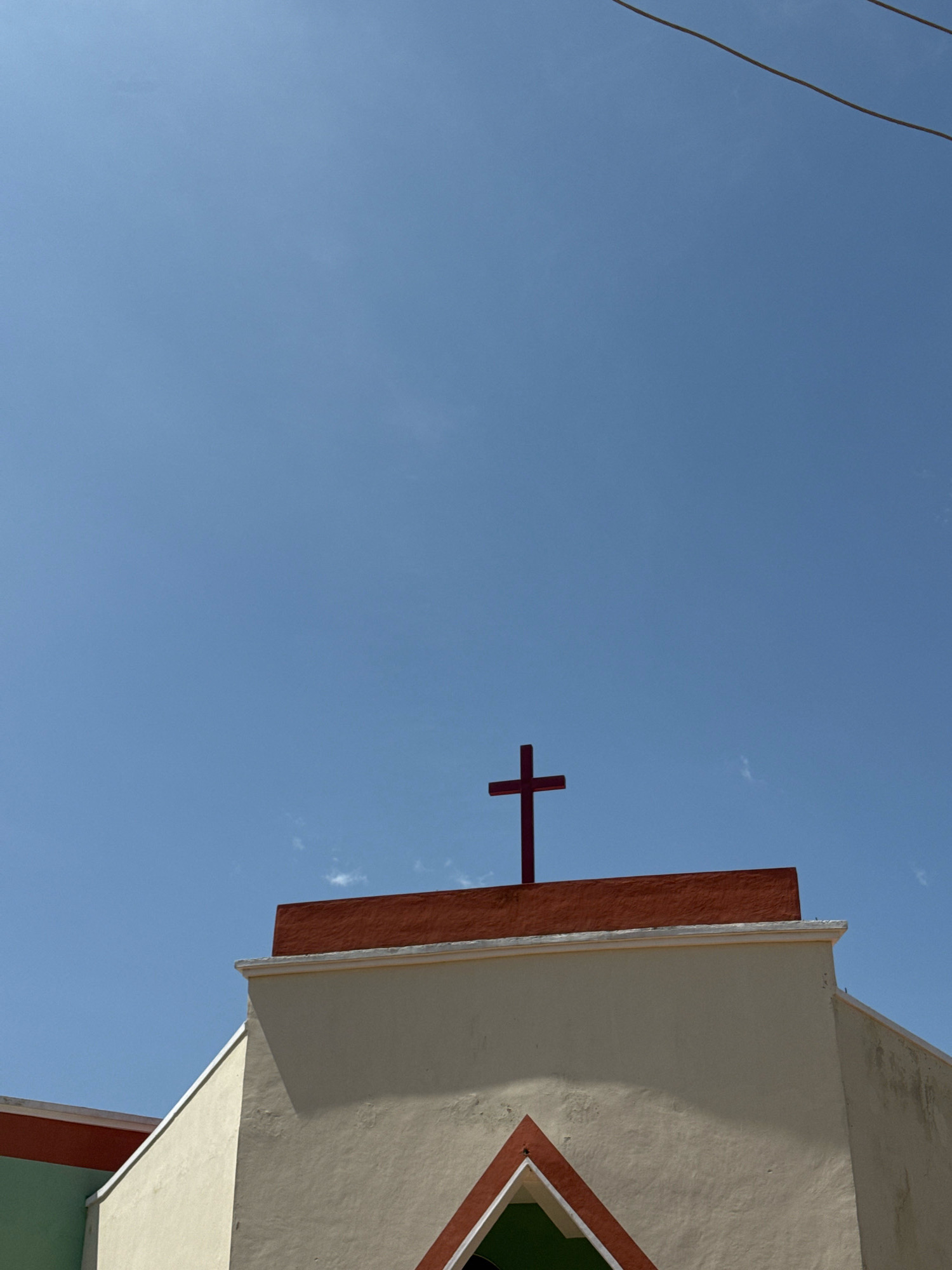 A close-up, low-angle shot captures the top of a building against a clear, pale blue sky. A dark red cross stands prominently on the building's rooftop, centered in the frame. The building's facade is painted a light cream color, with a reddish-brown trim along the top edge. A smaller section of the building on the left is painted green. The scene appears to be outdoors in a bright, sunny environment.