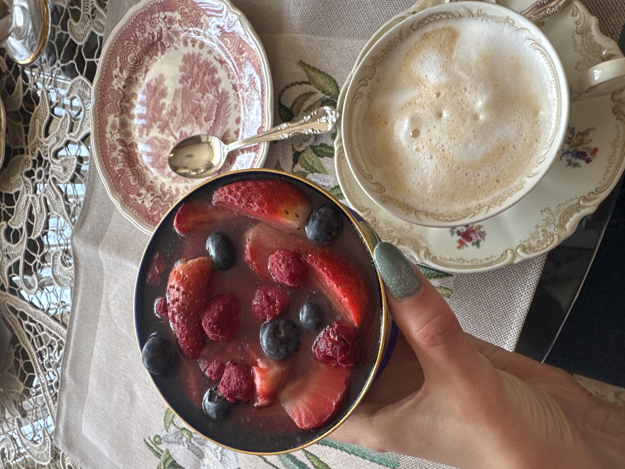 A high-angle shot captures a curated arrangement of food and drink on a patterned surface, suggesting a moment of leisure or a meal. In the lower center, a hand with a green, glittery nail holds a small, round bowl filled with a liquid and various berries, including sliced strawberries, raspberries, and blueberries.
To the left, a decorative plate with a red pattern sits with a silver spoon resting on it. To the right, a cup of frothy beverage is on a matching saucer. The background is a tablecloth with a botanical design that is partially obscured by a lacy surface in the upper left corner.
The location and city (Plovdiv, Bulgaria) are not discernible from the visual information provided. There are no people visible, beyond the hand. The primary subjects are the food and tableware.