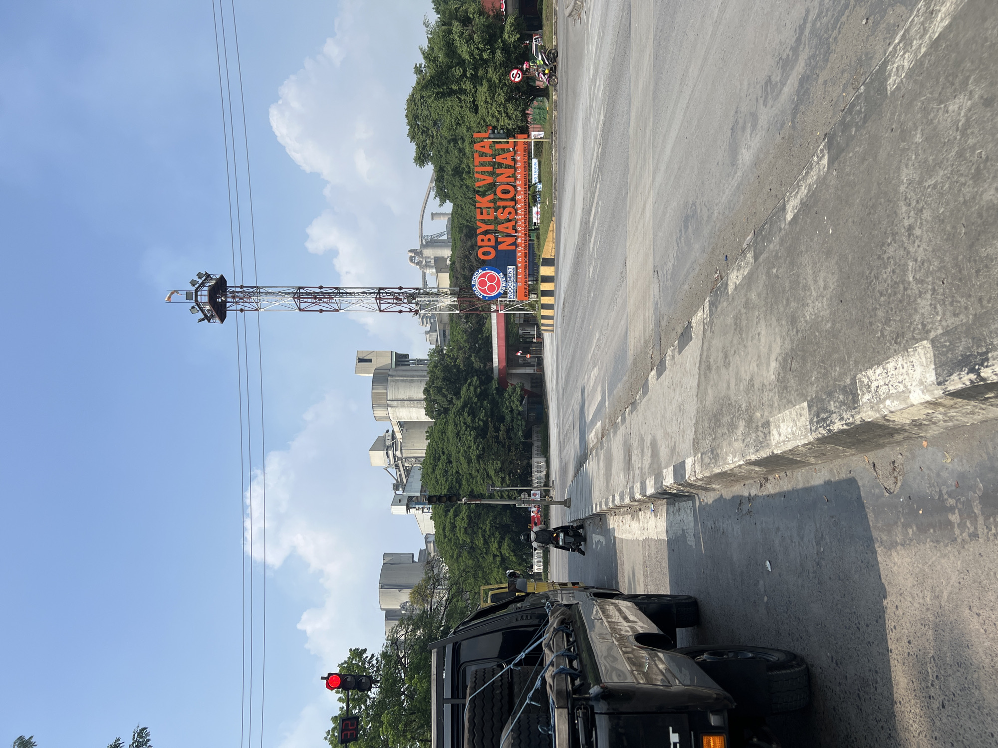 The image shows a street scene in Citeureup, Indonesia. In the foreground, a black pickup truck is parked on the left side of the road, loaded with tires. The road is paved and shows signs of wear and tear. To the right of the road, a sidewalk with black and white markings is visible. A traffic light is illuminated red, and a digital timer displays "32". A person on a motorcycle is stopped at the intersection.
In the background, a large industrial complex with several silo-like structures and a tall, lattice tower can be seen. A prominent sign displays "Obyek Vital Nasional" in large orange lettering, indicating a nationally vital object. The sky is blue with scattered clouds. Trees line the area between the road and the industrial buildings. Another traffic light is visible on the right side of the road, also illuminated red. A motorcycle with two riders is passing by on the right.