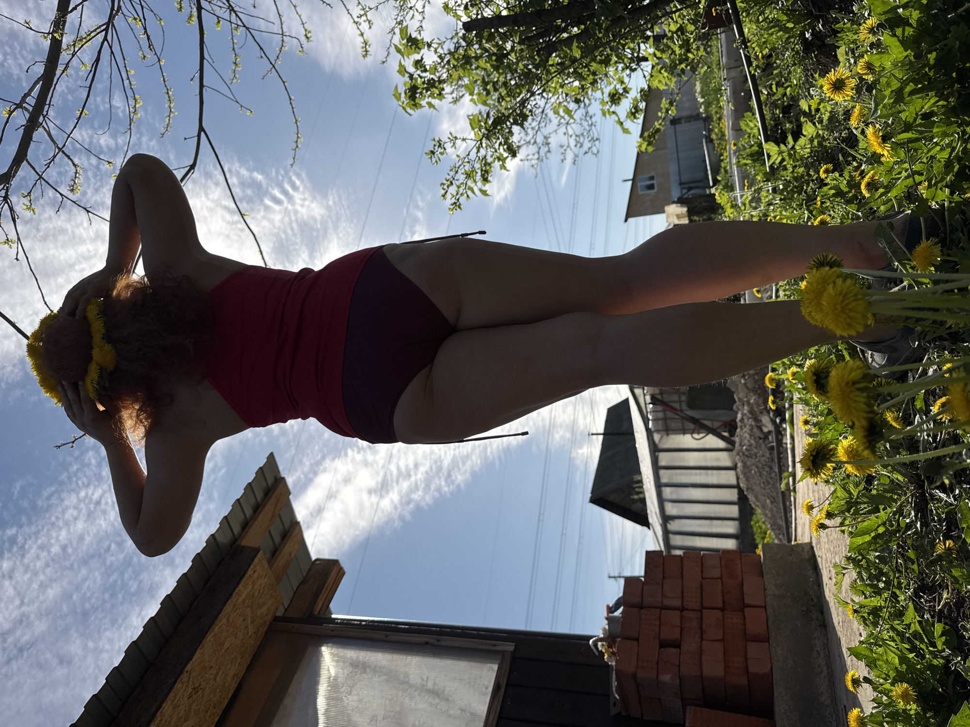 A woman with red hair is seen from a low angle wearing a red tank top and dark purple shorts. Her arms are raised towards her head. She is wearing a crown of yellow dandelions. She stands between a garden filled with dandelions and the side of a small building with a pile of bricks nearby. Overhead are power lines, foliage, and a partially cloudy sky. A house is visible in the background. The location is Chaikovsky, Russia.