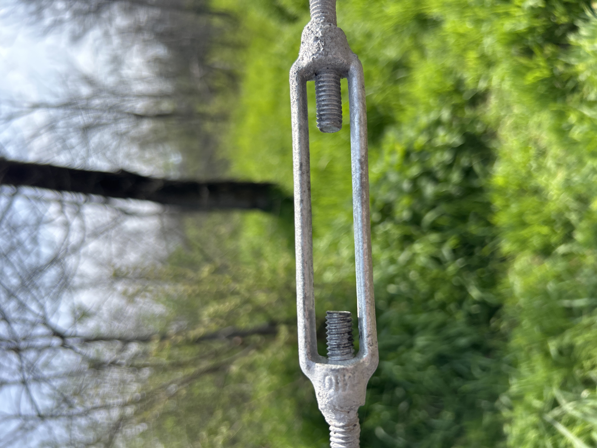 The image shows a close-up of a metal turnbuckle tensioner against a blurred background. The turnbuckle is silver in color and displays threaded ends. The background is split into two distinct areas: on one side, blurred green foliage suggests a grassy or wooded area; on the other side, a similar blurred scene suggests trees without leaves, possibly with a body of water beyond. It is not possible to determine the specific location within Plovdiv, Bulgaria, without additional information or landmarks. There are no people visible in the image.